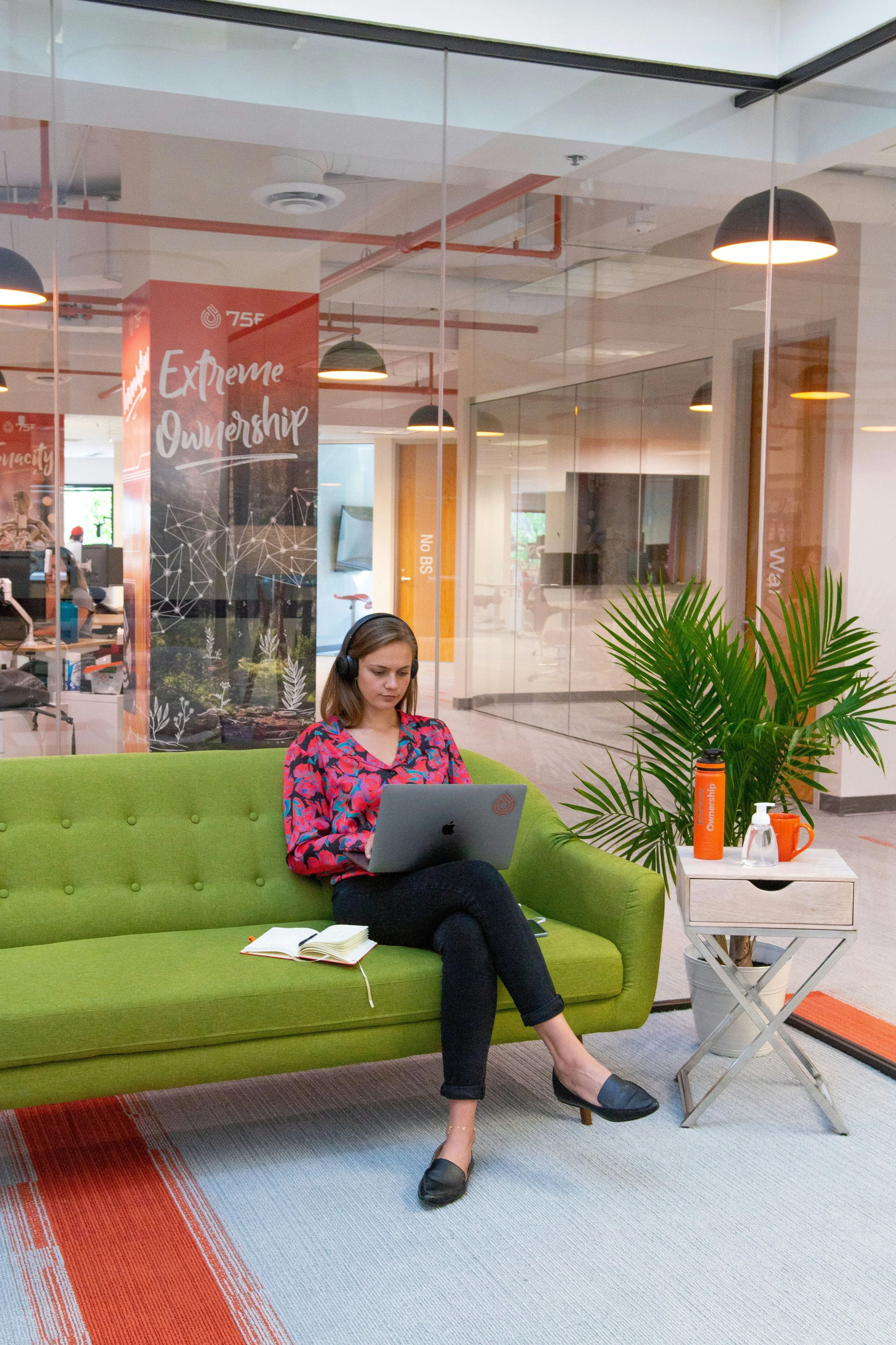 Woman sitting on a green sofa with a laptop, wearing headphones, in a modern office space with glass walls and plants.