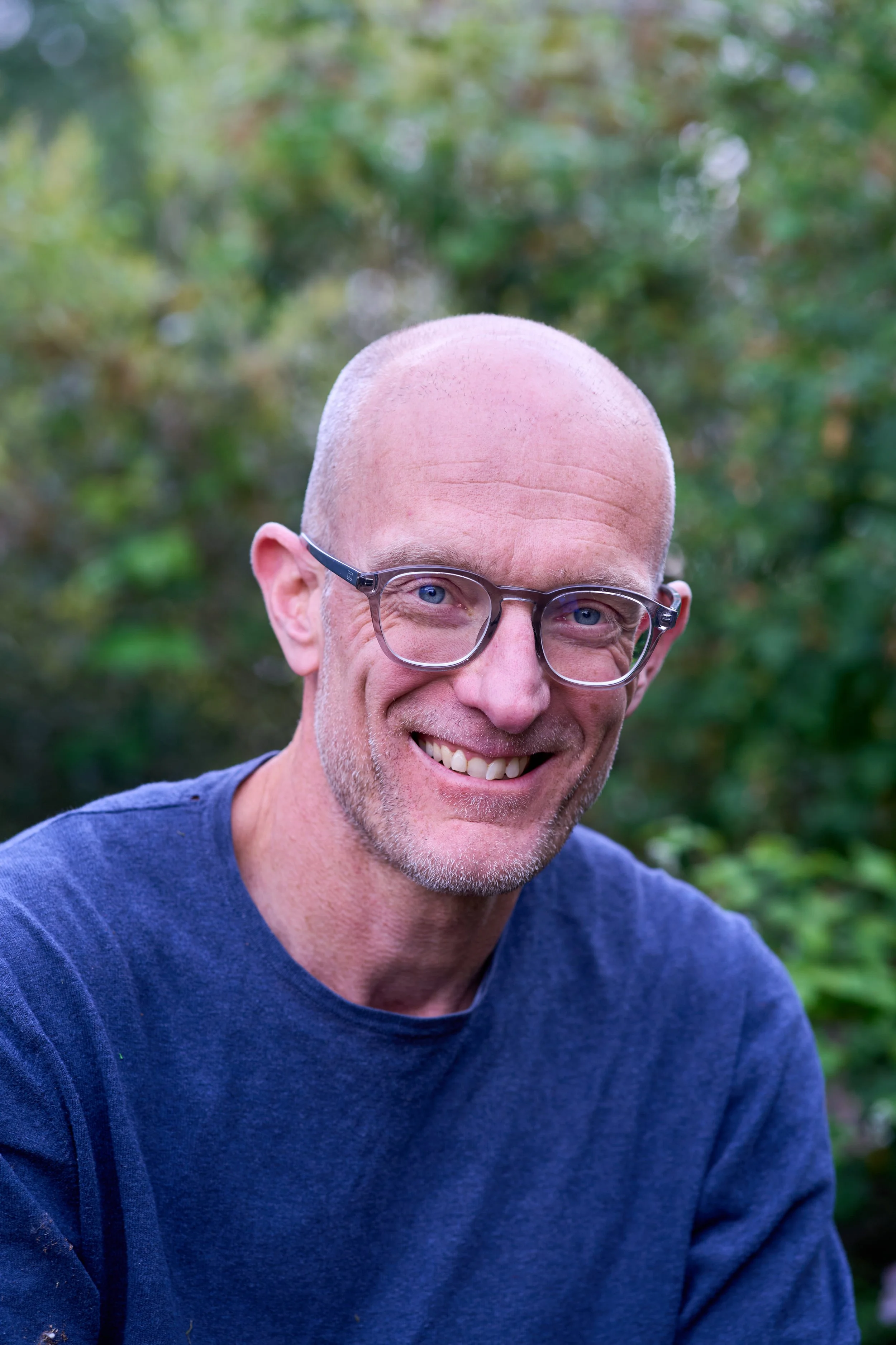 Smiling bald man with glasses outdoors in a green, blurry background.