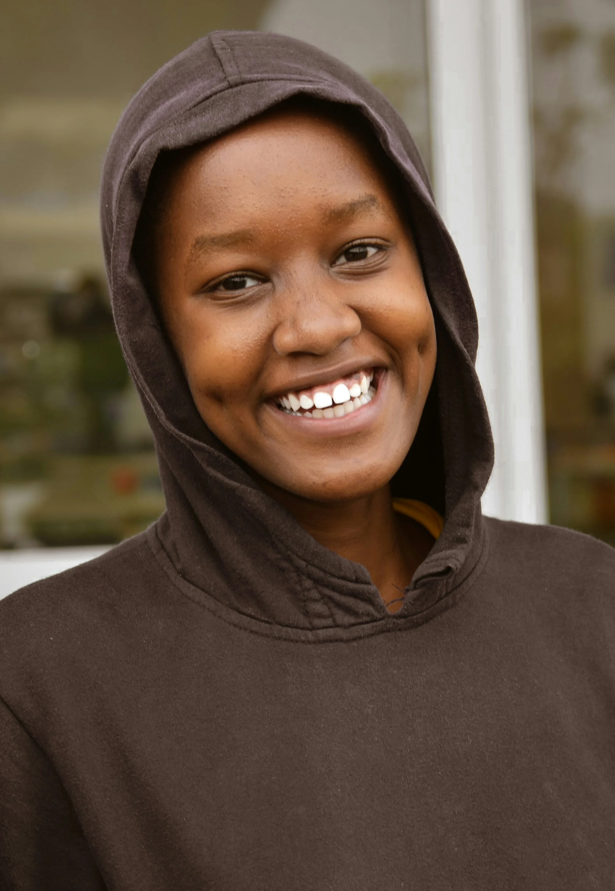 A smiling young woman wearing a dark hoodie standing outdoors in front of a window.