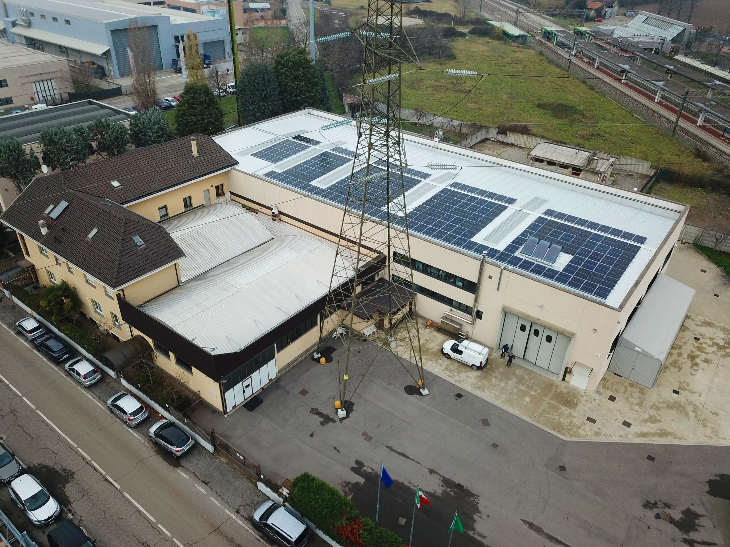 Edificio industrial con pannelli solari sul tetto, situato vicino a una strada con automobili parcheggiate. È presente anche una linea elettrica alta con un traliccio.