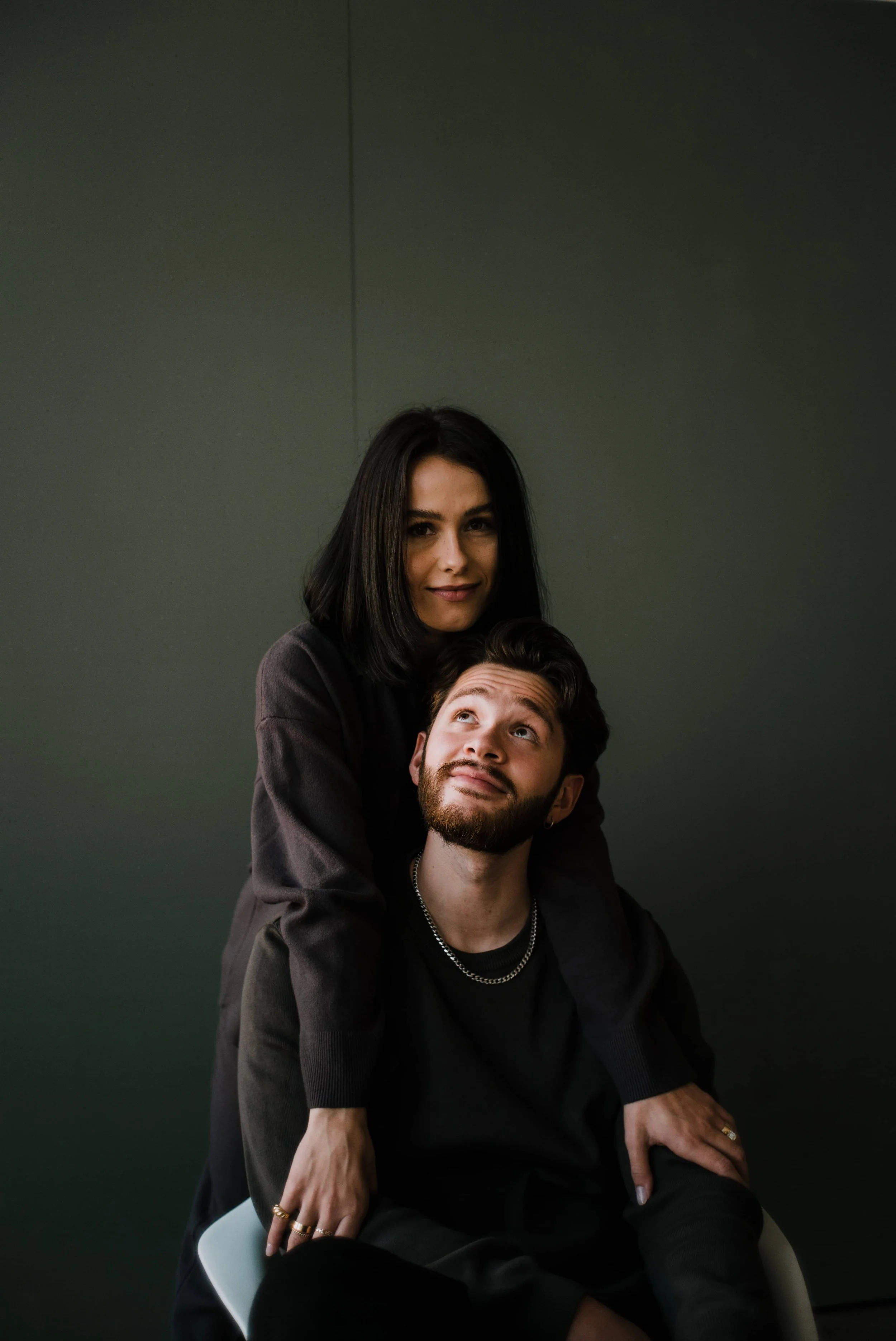 A woman with black hair and a man with dark hair and a beard sitting and leaning on each other against a plain dark background.