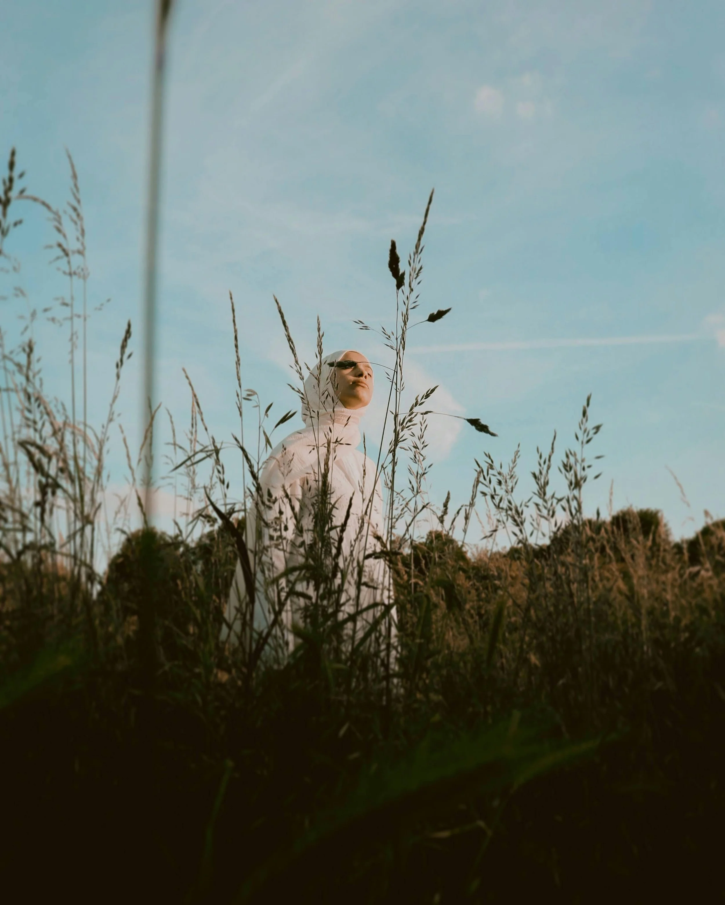 Low angle view of a woman with a headscarf and closed eyes standing among tall grasses in a field during sunset.