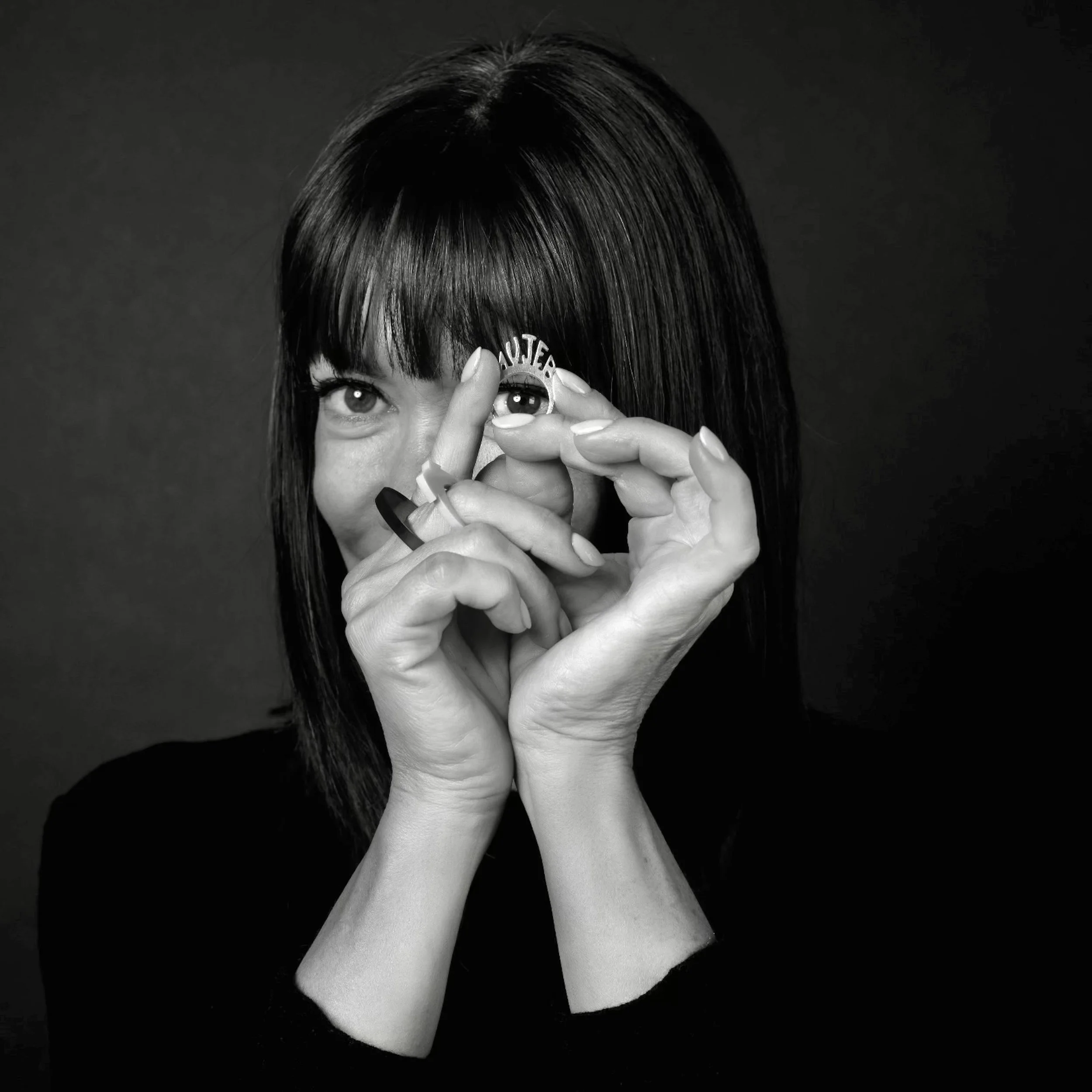 A woman with dark hair and bangs peeks through her fingers, holding a ring upper her eyes looking throuth, against a dark background.
