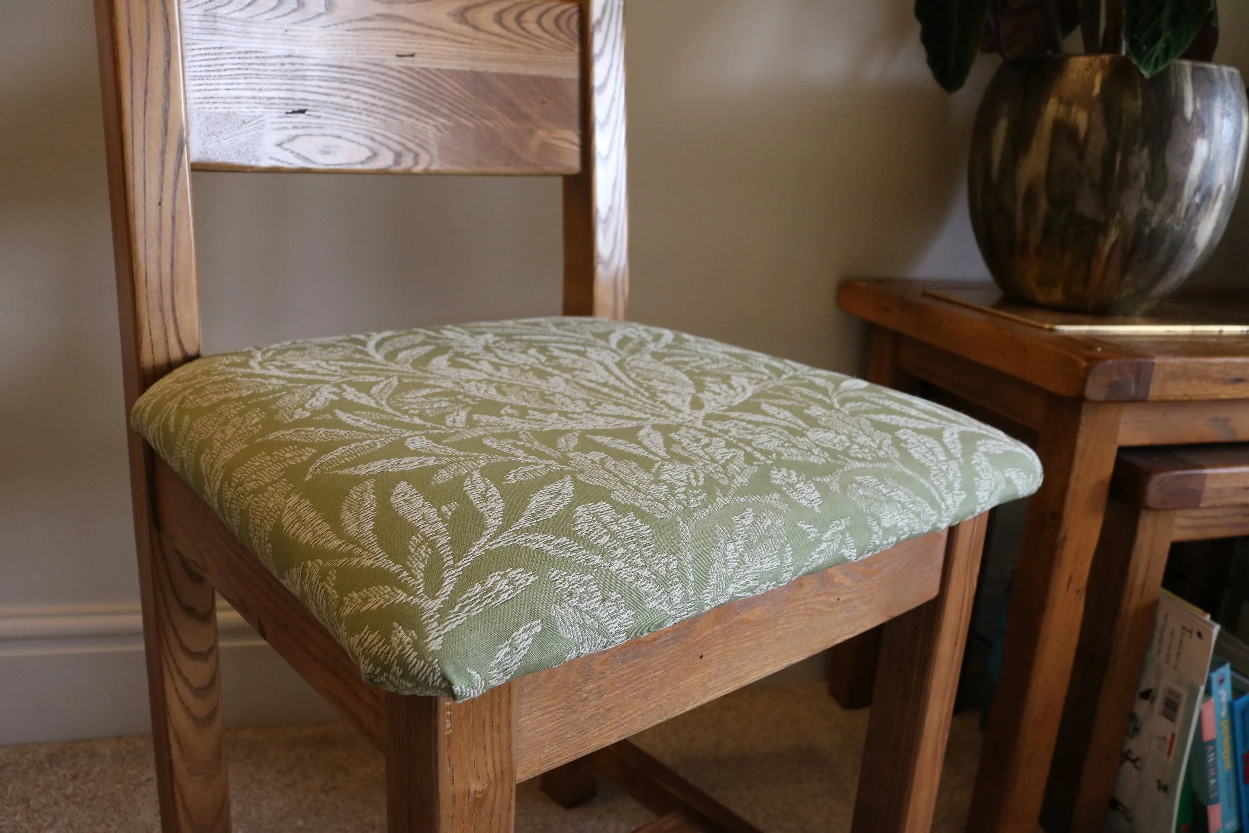 A wooden chair with a green padded cushion featuring a white leafy pattern, positioned next to a wooden side table with a large decorative vase and a potted plant on top.