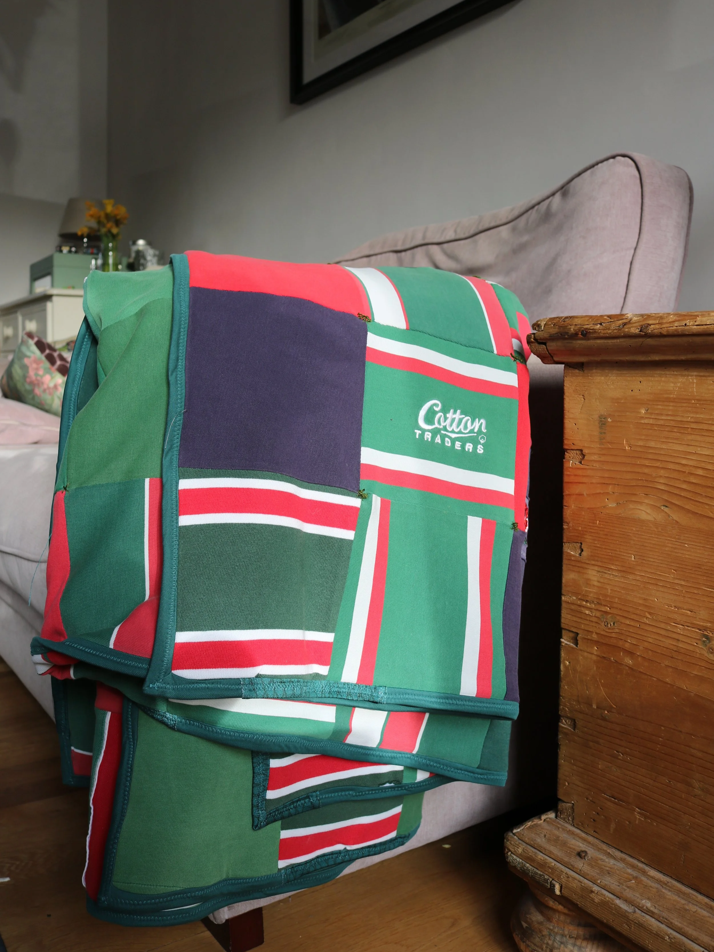 A colorful quilted blanket with red, green, white, and navy stripes and blocks resting on a beige sofa, beside a wooden side table, in a cozy living room.