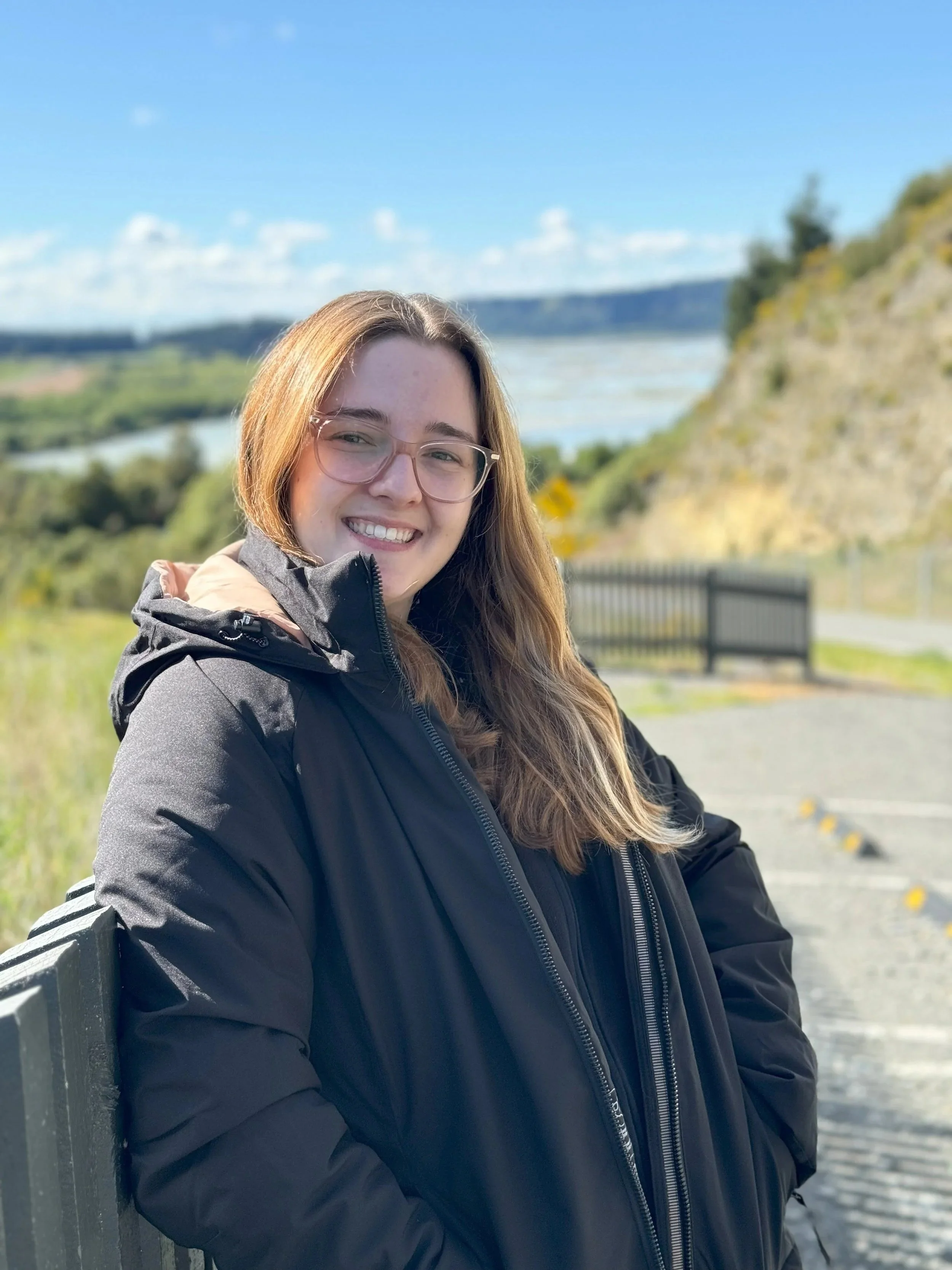 A young woman with long light brown hair, glasses, and a smiling expression, wearing a black jacket, stands outdoors near a body of water and a hill, with a partly cloudy sky in the background.