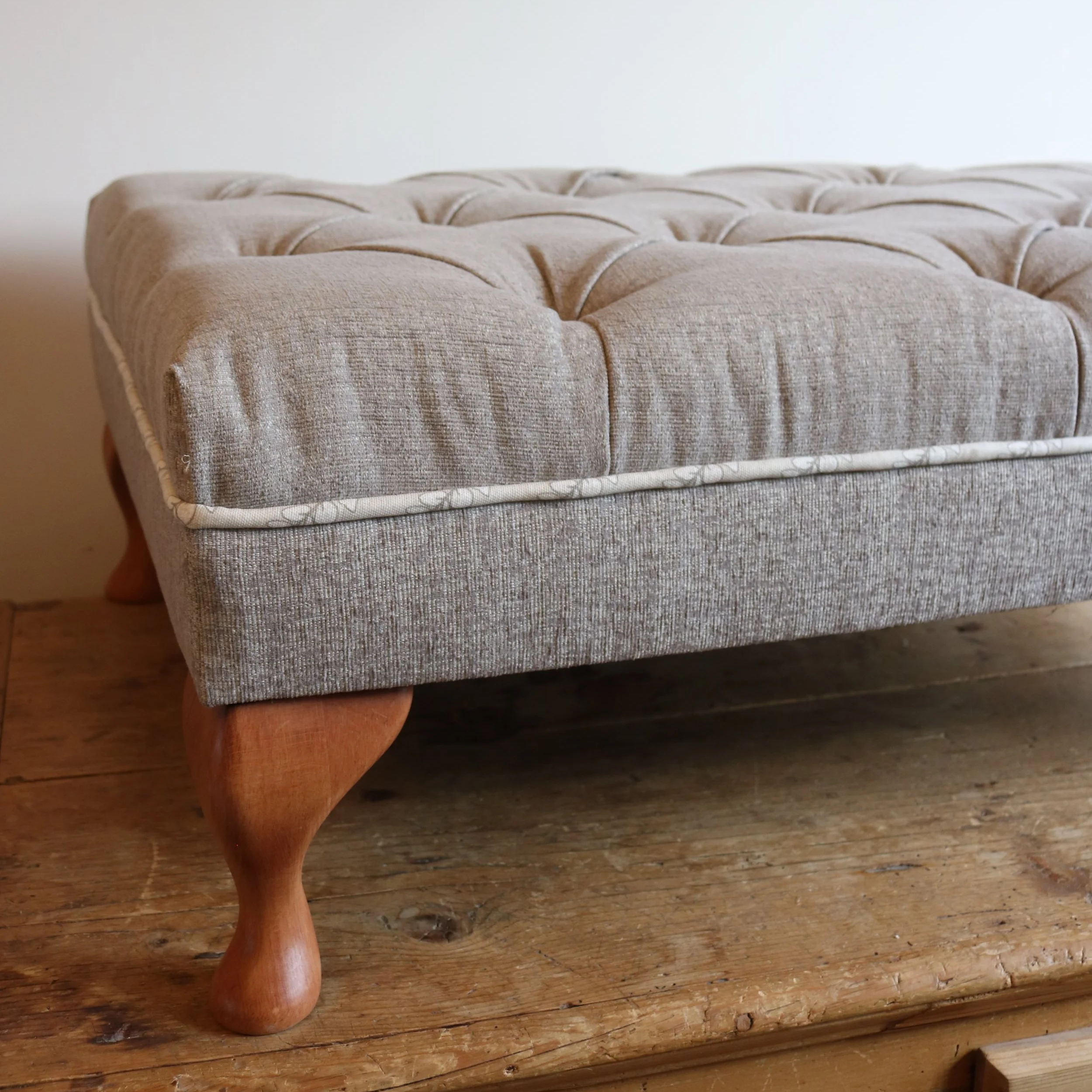 Close-up of a beige upholstered ottoman with wooden cabriole legs, sitting on a wooden floor.