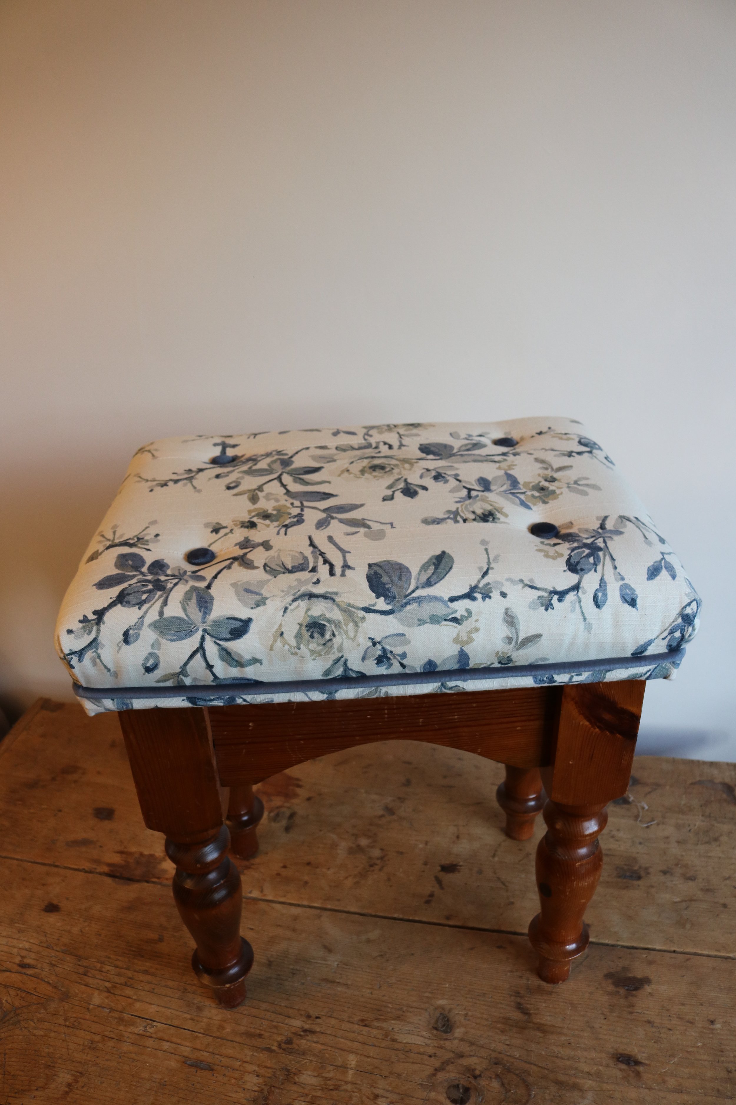 A small wooden stool with turned legs and a cushioned top, placed on a wooden surface against a plain wall.
