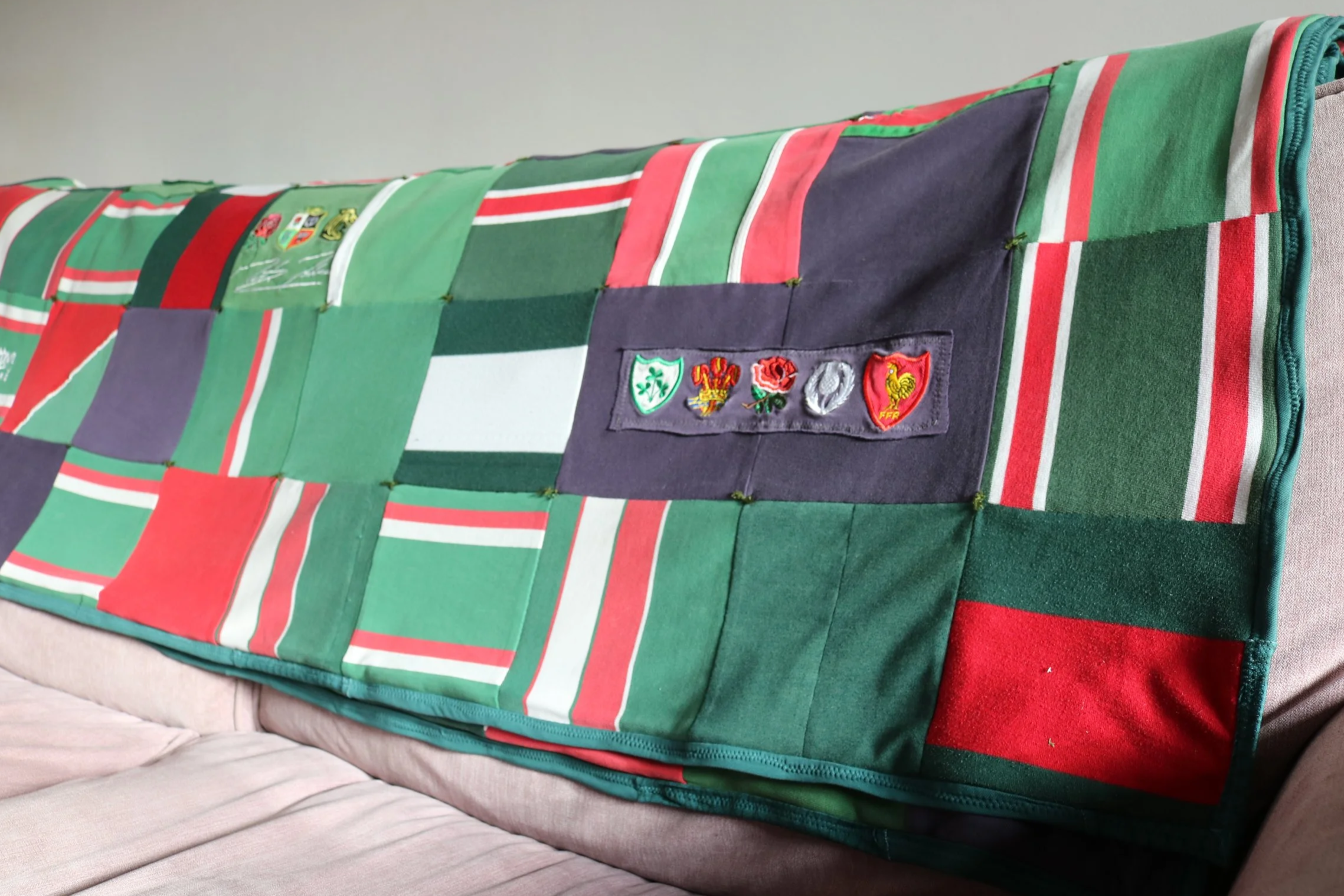 Colorful quilt made of patchwork fabric squares in red, green, white, purple, and gray, draped over a couch. The quilt features a row of embroidered emblems or badges including a green shield, a red and yellow rooster, and other symbols.