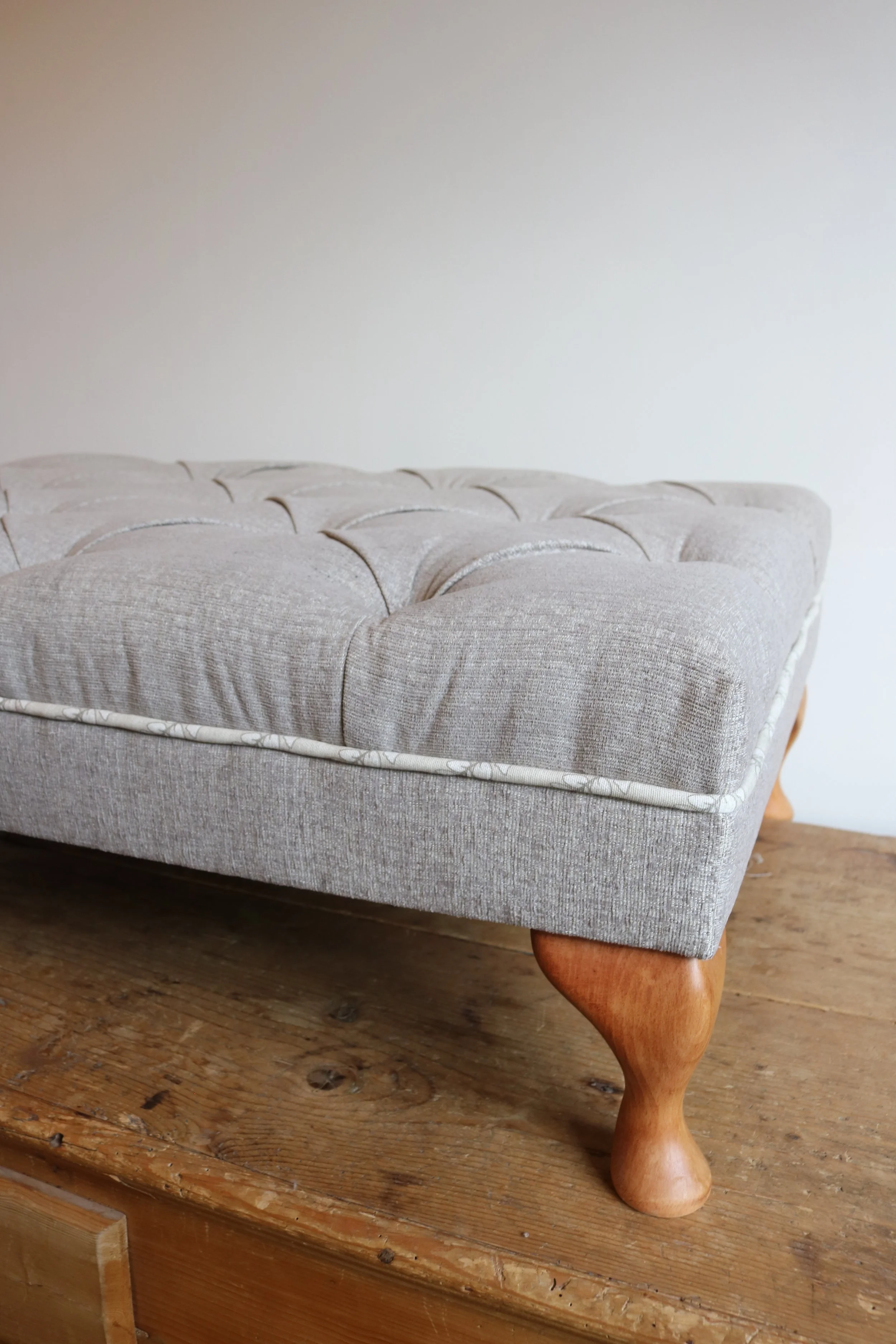 Close-up of a tufted beige upholstered furniture piece with a wooden leg on a wooden surface against a plain wall.