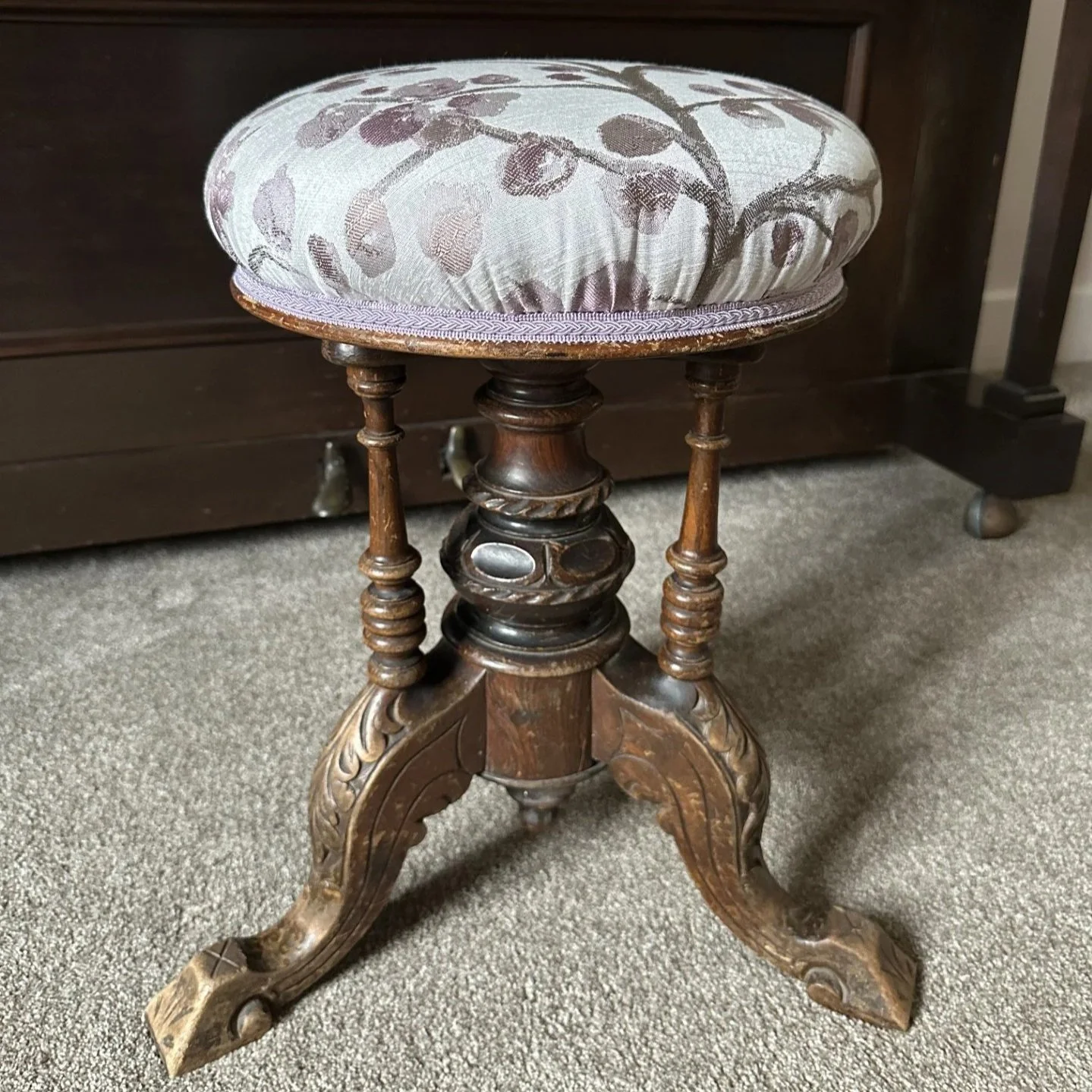 An antique wooden stool with a padded, floral-patterned fabric cushion on top, placed on a carpeted floor.