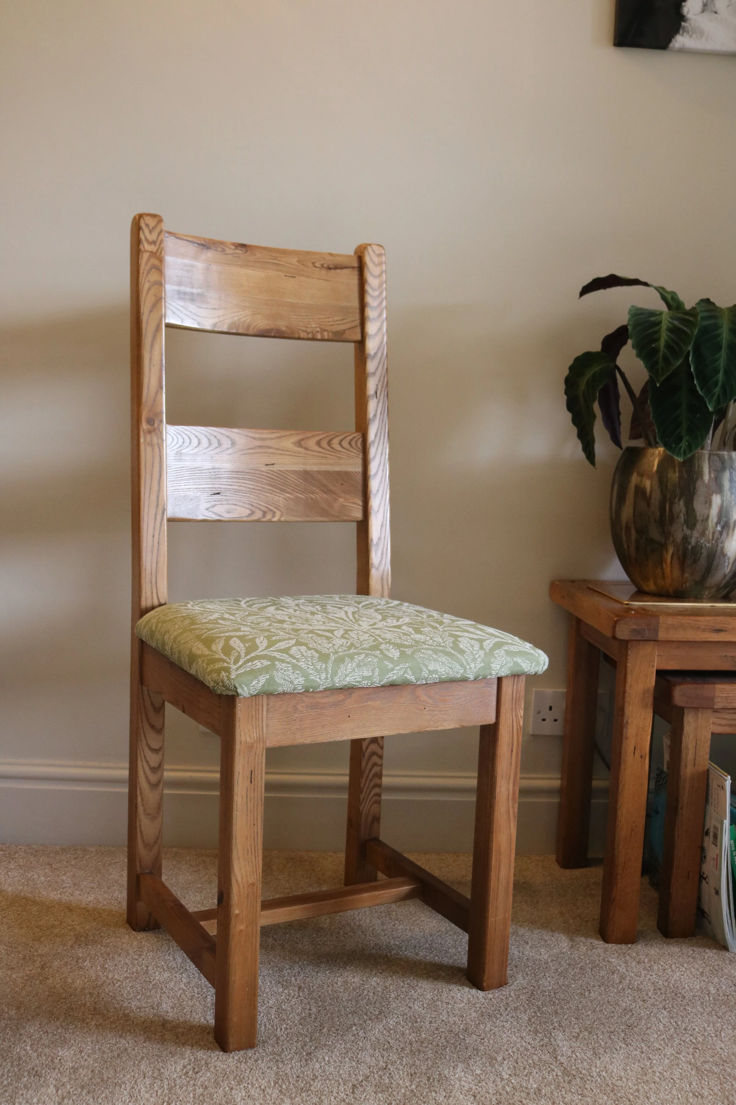 A wooden chair with a green patterned cushion next to a wooden side table holding a large plant in a metallic pot.