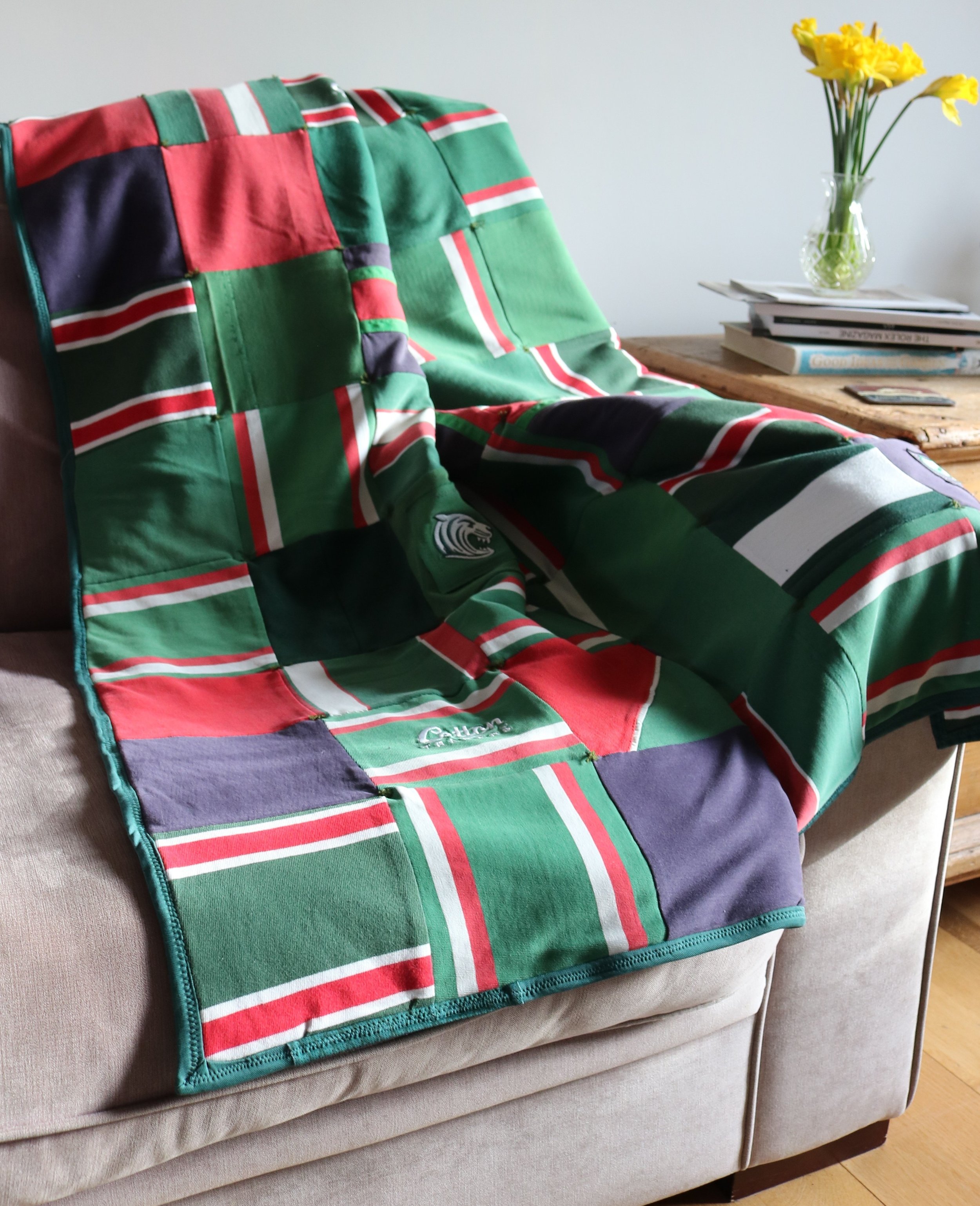 Colorful quilt with green, red, white, and purple checks draped over a beige sofa, with a wooden side table holding a vase of yellow flowers, books, and a remote control.