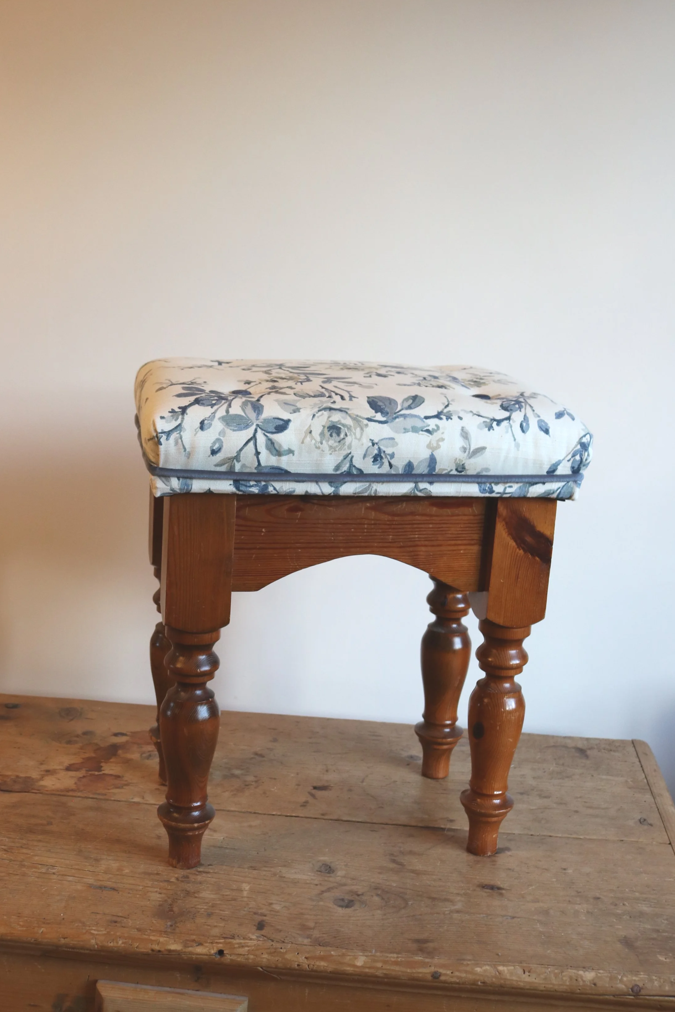 A wooden stool with turned legs and a cushioned seat with a floral pattern, placed on a rustic wooden surface.