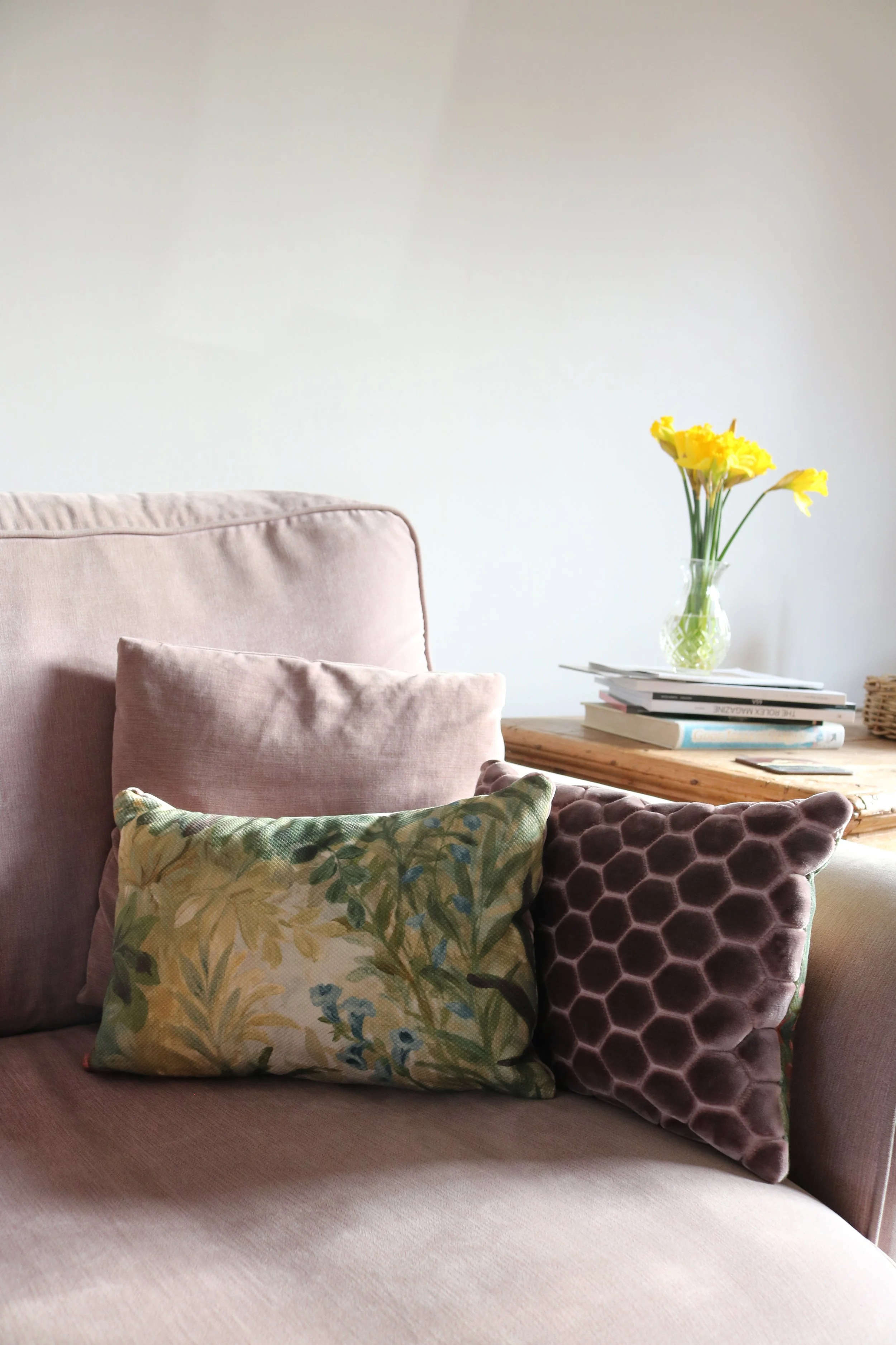 A beige sofa with three decorative cushions: one pink velvet, one floral patterned, and one purple velvet with a honeycomb pattern; a wooden table with a vase of yellow flowers and some books in a well-lit room.