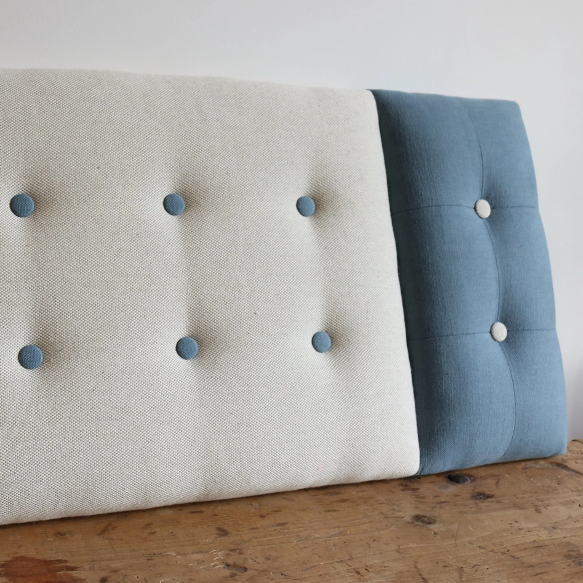 Close-up of a fabric headboard with button tufting, featuring a beige section on the left and a blue section on the right, resting on a wooden surface.