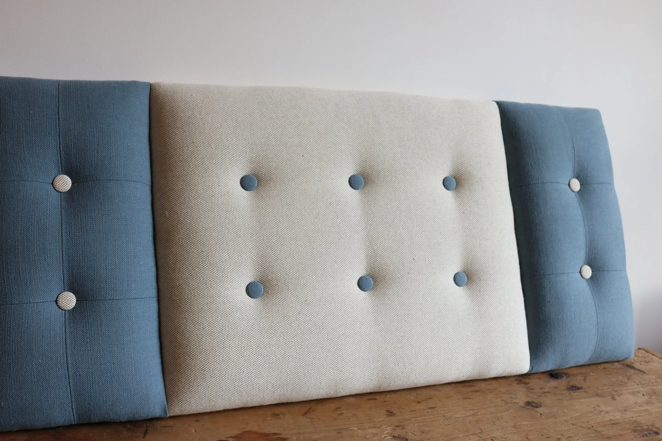 Close-up of a padded headboard or panel with three sections: blue, beige, and blue, each with tufted buttons, mounted on a wooden surface against a white wall.