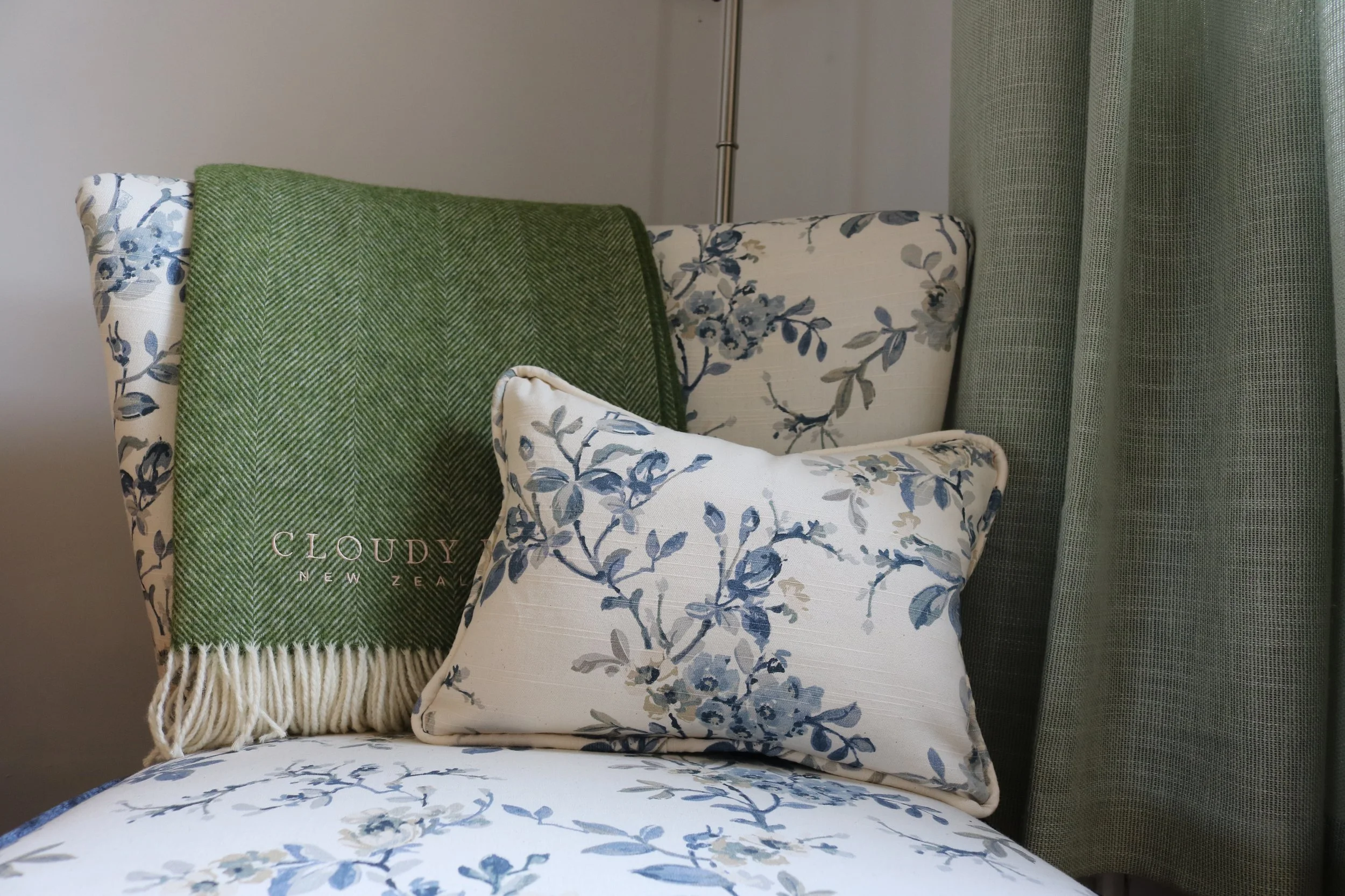Close-up of floral-patterned cushions and a green woven blanket on a chair next to a window with green curtains.
