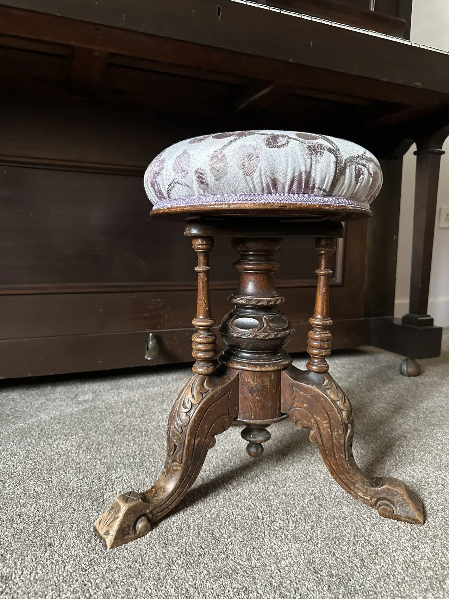 A vintage wooden stool with ornately carved legs and a padded, upholstered seat in a fabric with a floral pattern, placed on a carpeted floor.