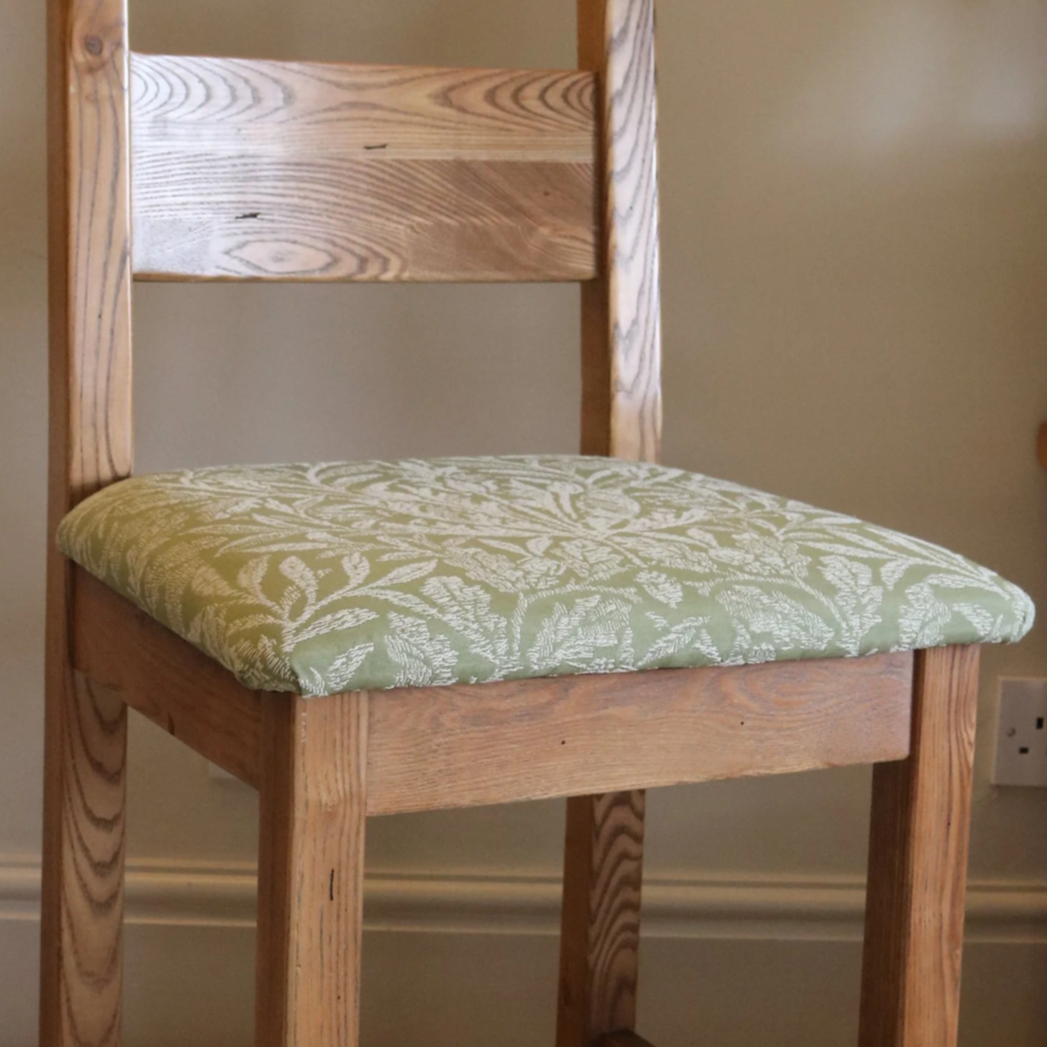 A wooden chair with a cushioned seat upholstered in green fabric with a white floral pattern, positioned against a beige wall near an electrical outlet.