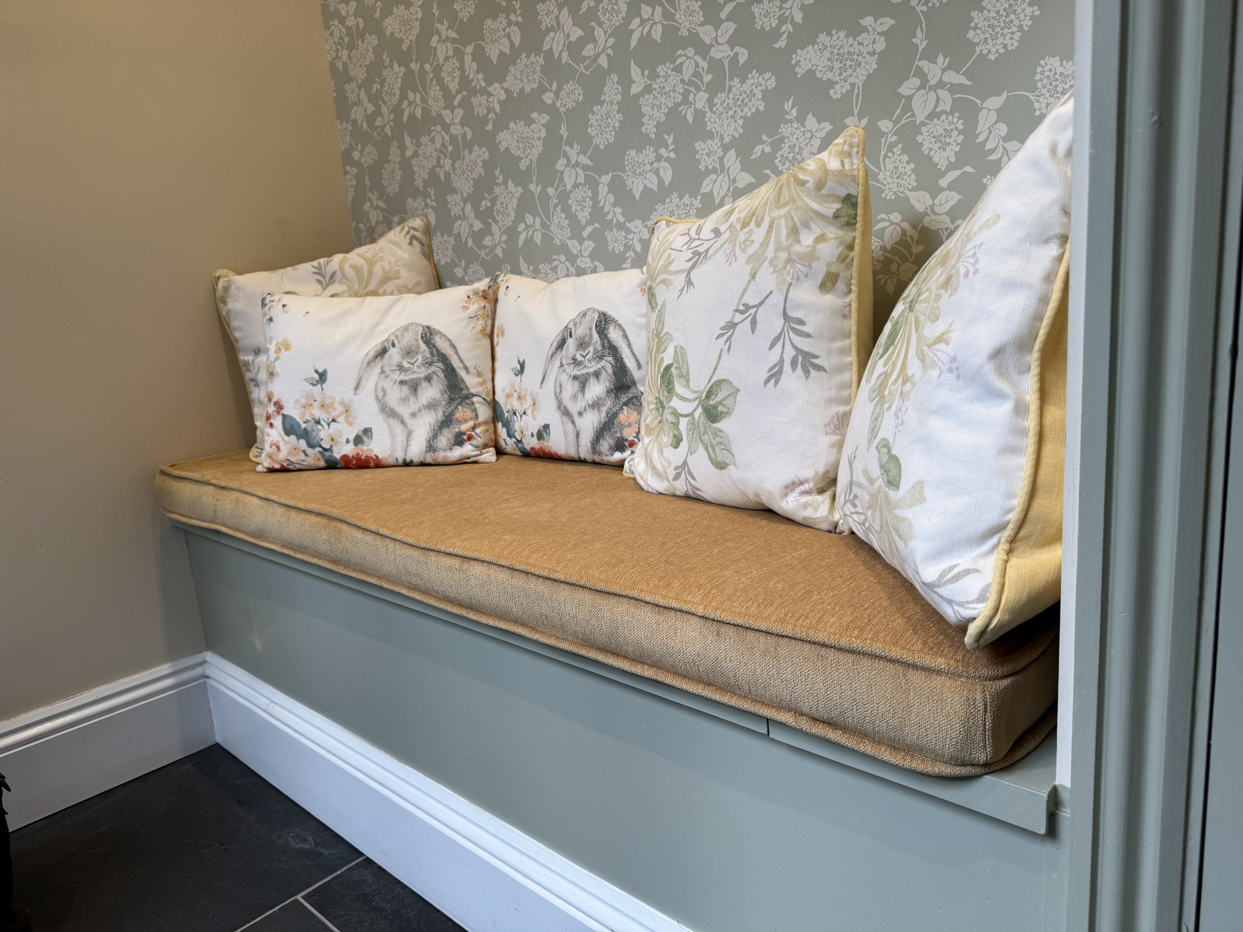 A built-in window seat with a beige cushion and multiple decorative pillows featuring floral and bunny designs, against a wall with floral patterned wallpaper and a beige wall.