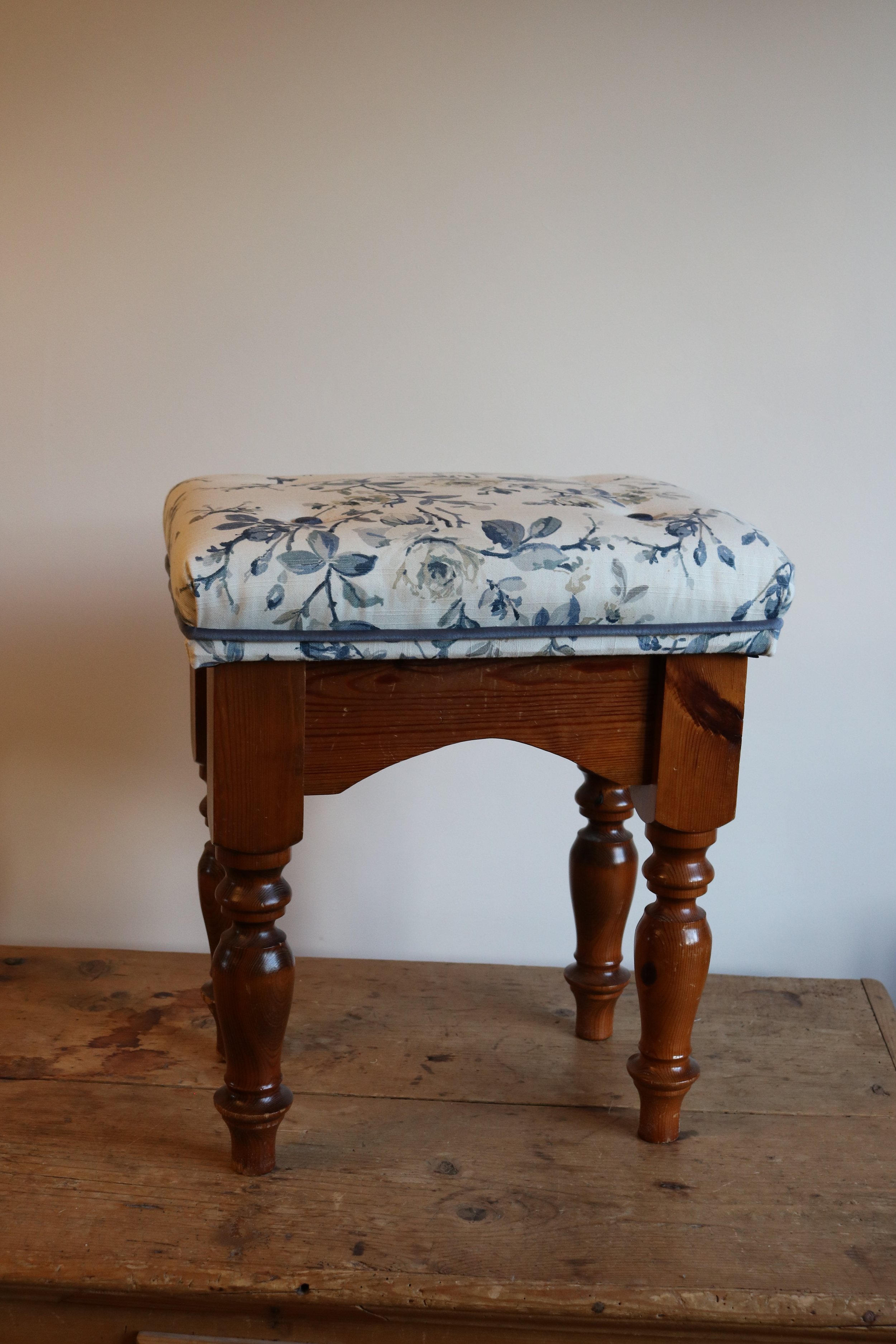 A small wooden stool with turned legs, topped with a cushioned seat covered in floral fabric, placed on a worn wooden surface against a plain wall.