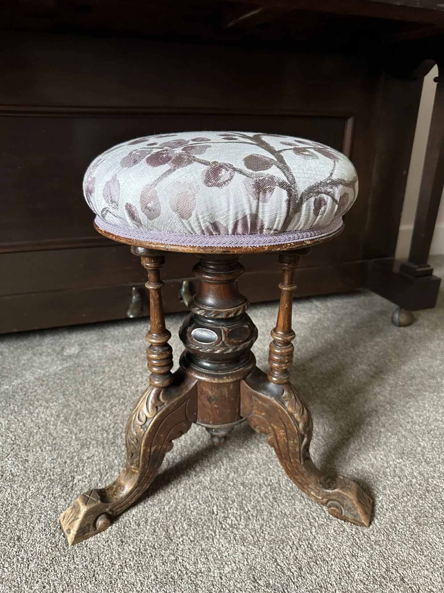 An antique wooden stool with a padded fabric cushion on top, featuring a floral pattern, placed on a carpeted floor in front of a dark wooden piece of furniture.