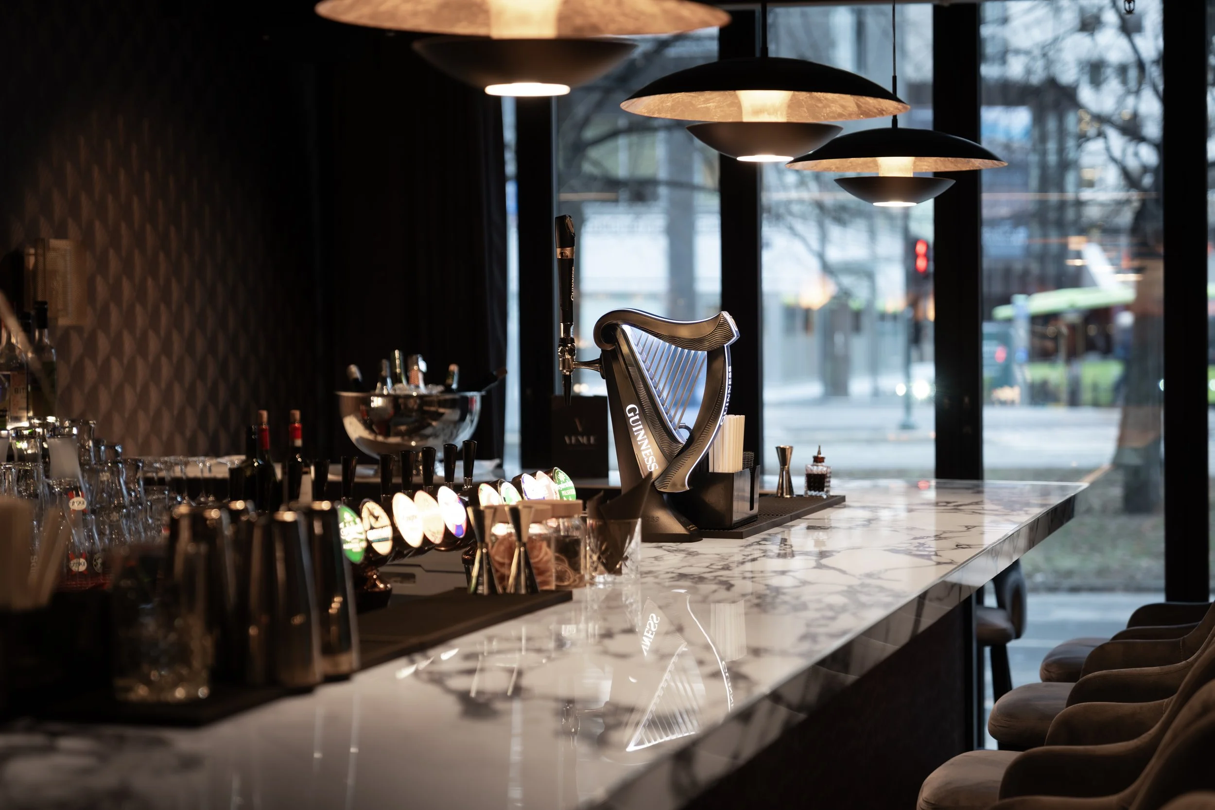 Interiør av en bar med en marmorben, lysende øl-pressere, en harpespill for å lage drinker, og glass foran. Store vinduer viser en utendørs gate og trær.