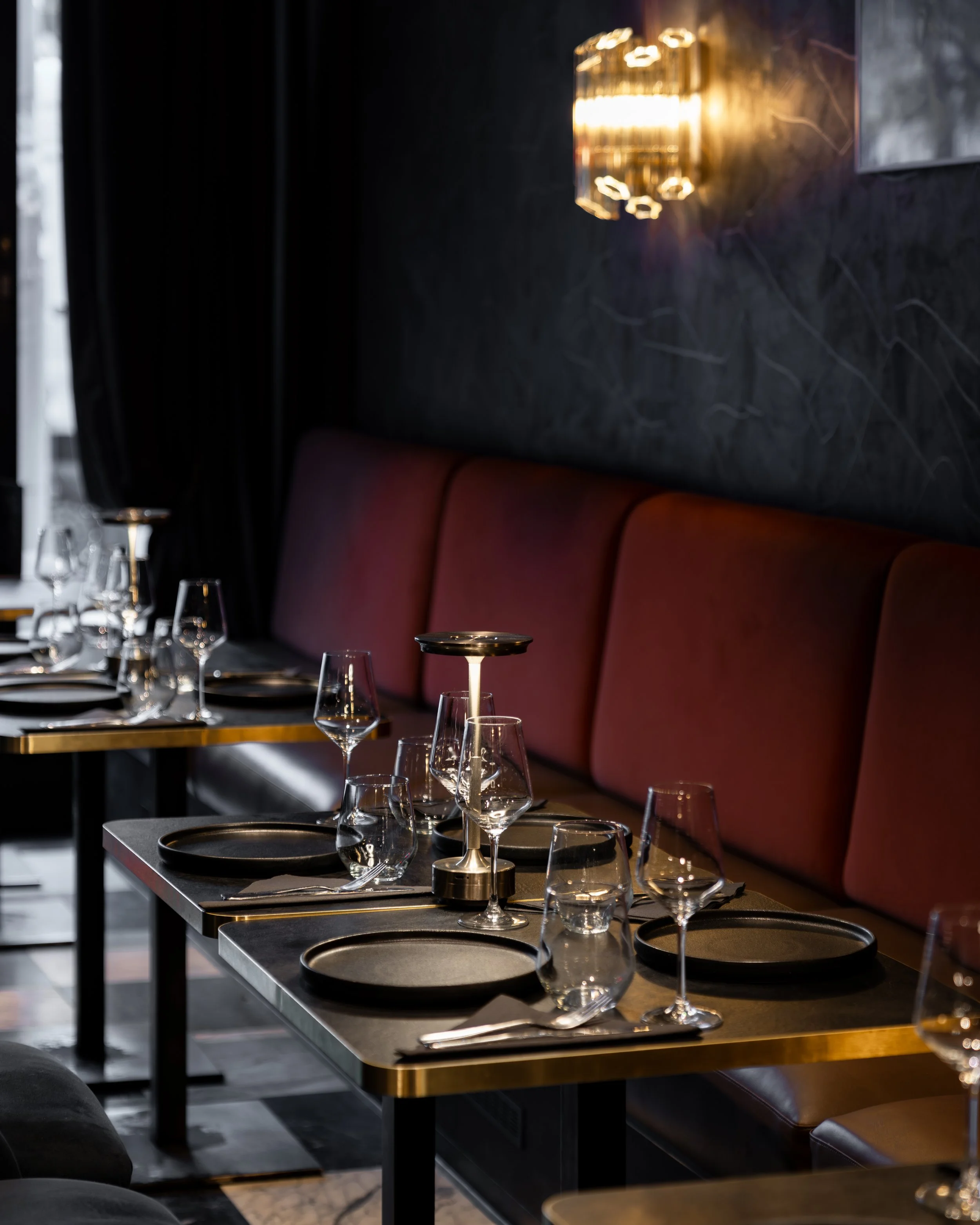 Elegant restaurant bord dekket med svarte tallerkener, tomme vinglass og sølvbestikk, med en dekorativ lykt i midten, omgitt av myk belysning og mørke veggdekorasjoner. Sofaen er av luksuriøst fløyel eller velour materiale. 