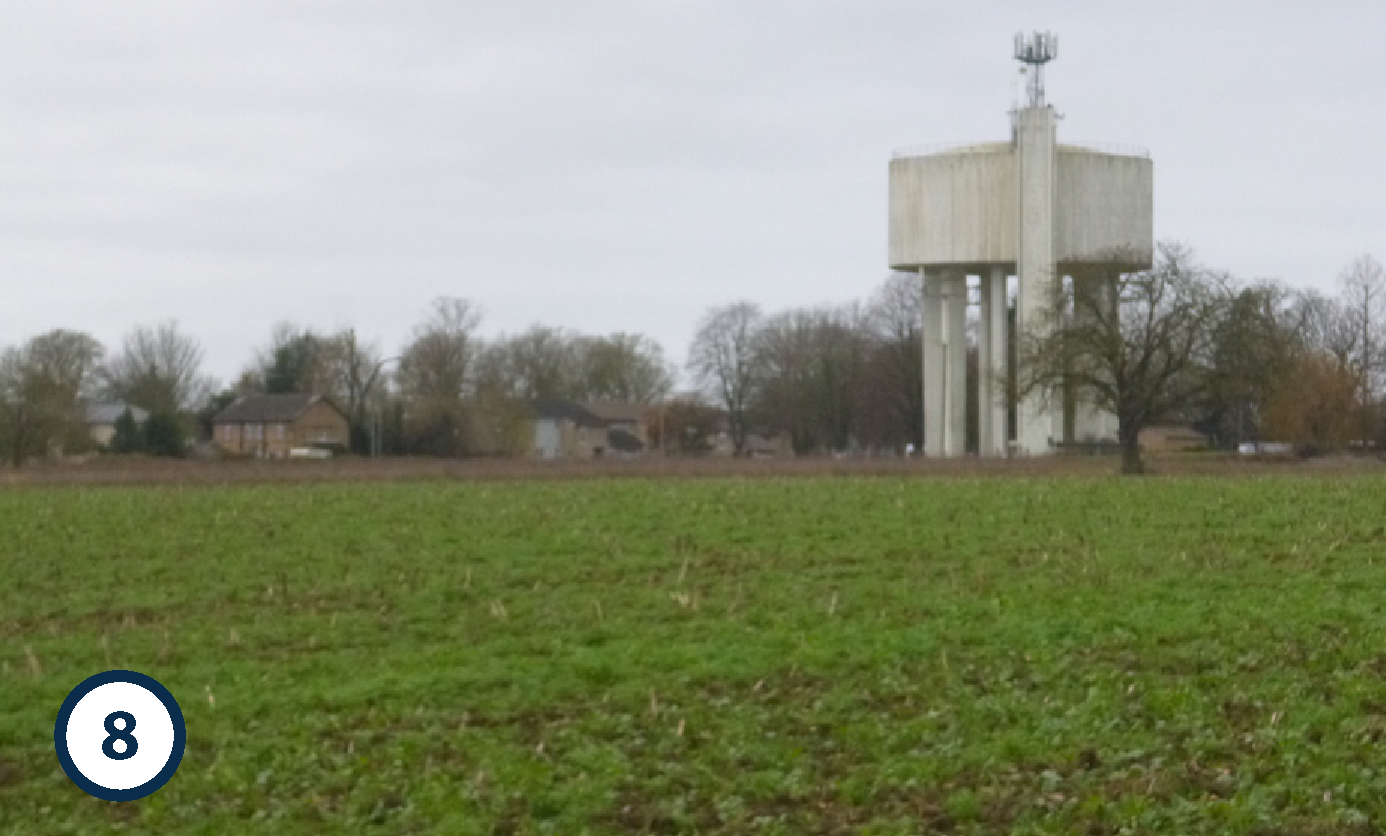 A water tower standing in a rural area with trees and houses in the background under an overcast sky.