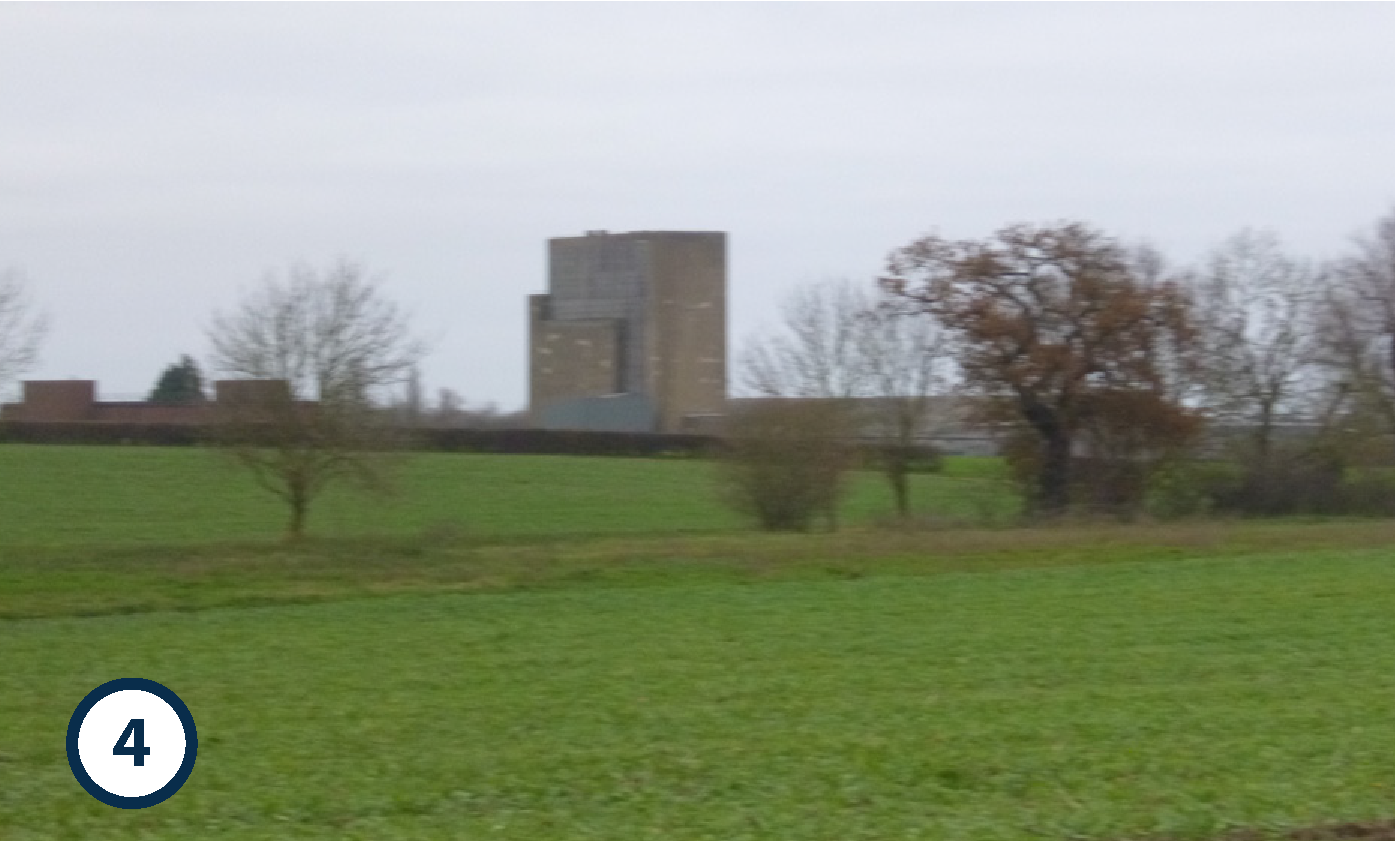 Blurred image of a grassy field with trees and a large concrete building in the background under an overcast sky.