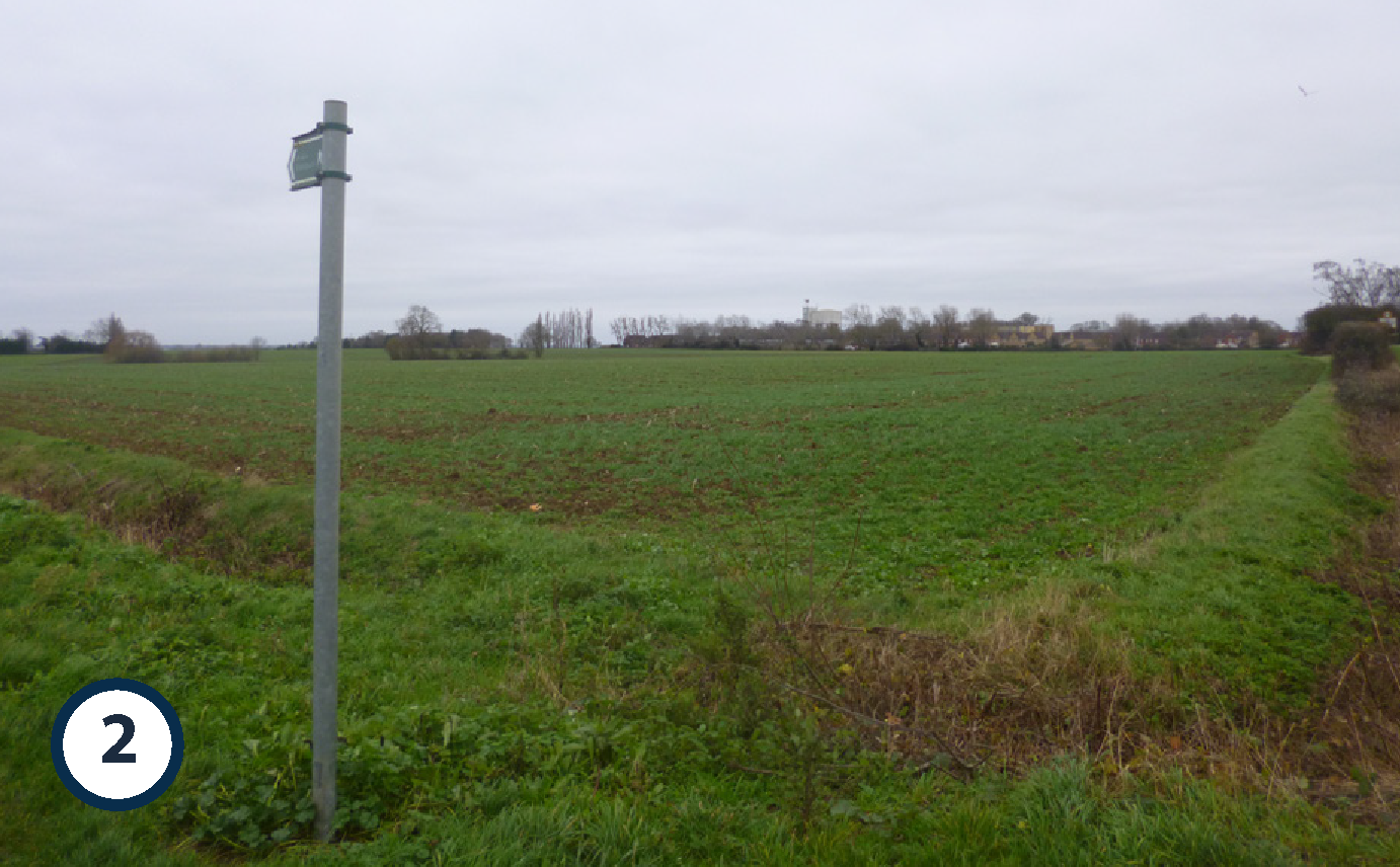 A green agricultural field under a cloudy sky with a metal pole on the left and a rural landscape in the distance.