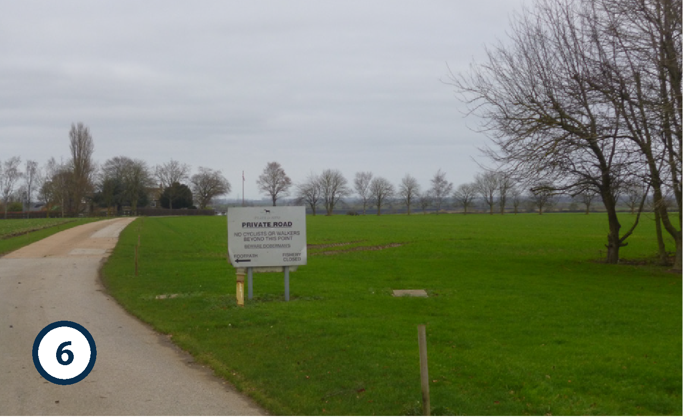 A rural scene with a gravel road curving to the left, bordered by green grass and leafless trees against a cloudy sky. There is a white sign indicating a private road with restrictions and closures.