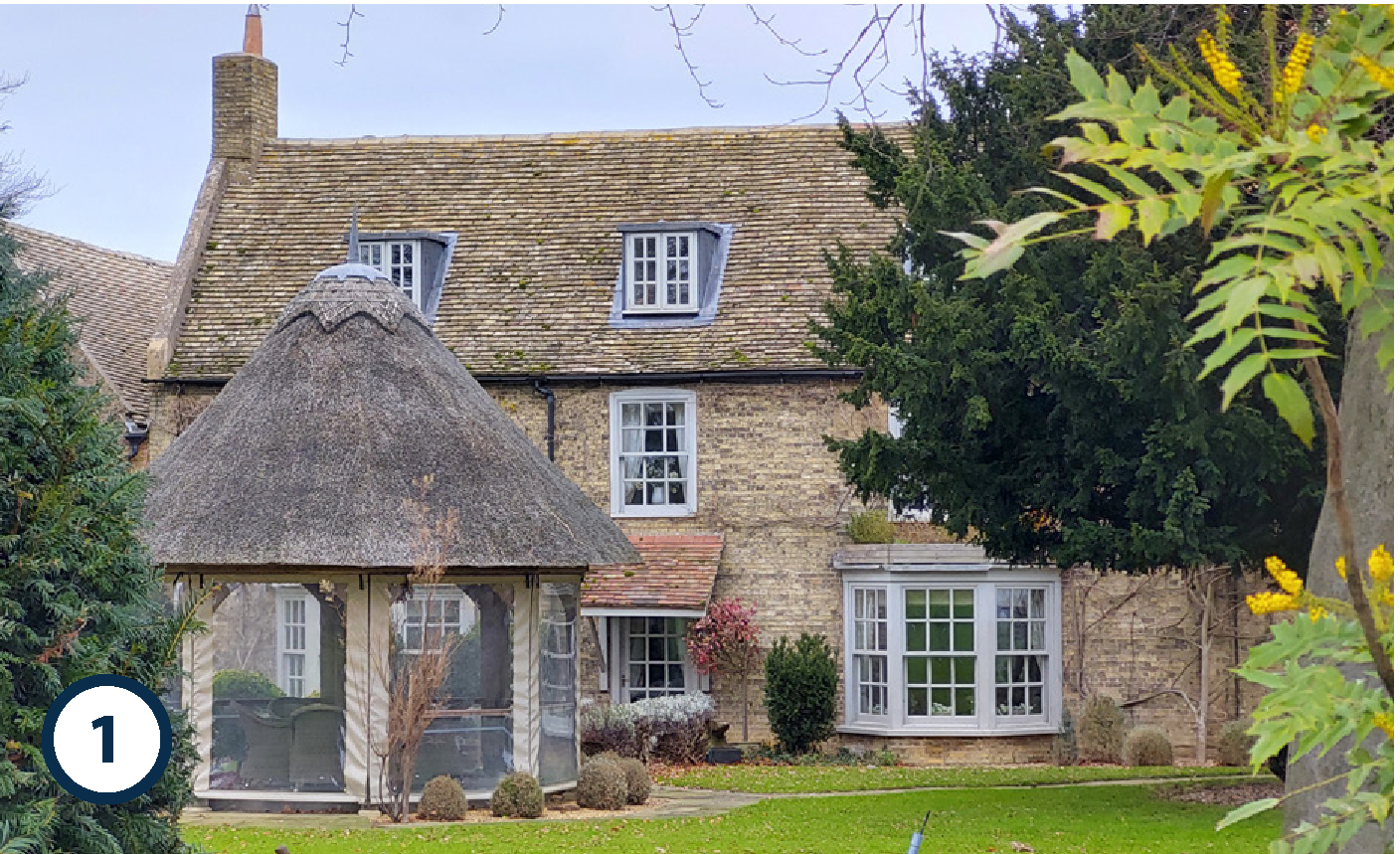 A stone house with a sloped roof featuring small dormer windows. In front, there is a gazebo with a thatched roof and mesh sides, surrounded by a well-kept lawn, bushes, and trees.