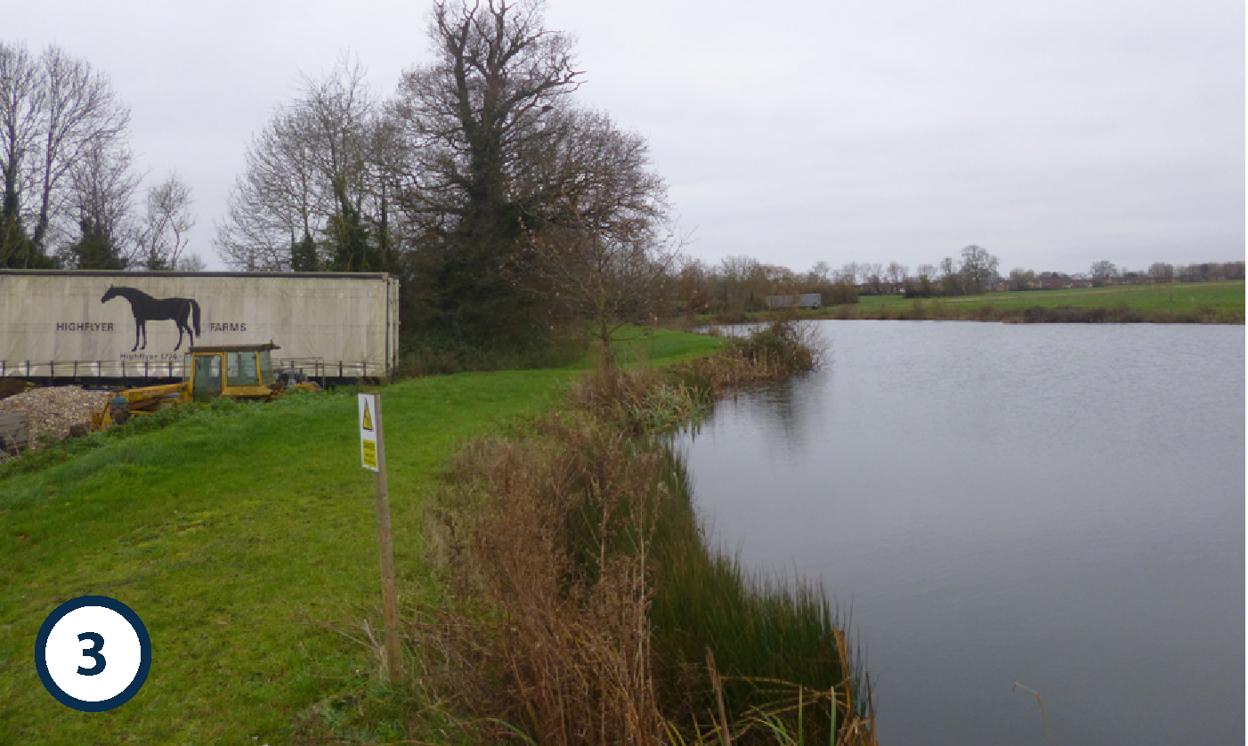 A rural scene with a canal on the right, grassy banks, leafless trees, and an industrial trailer with a horse logo and the words 'Highflyer Farms' in the background. Overcast sky.
