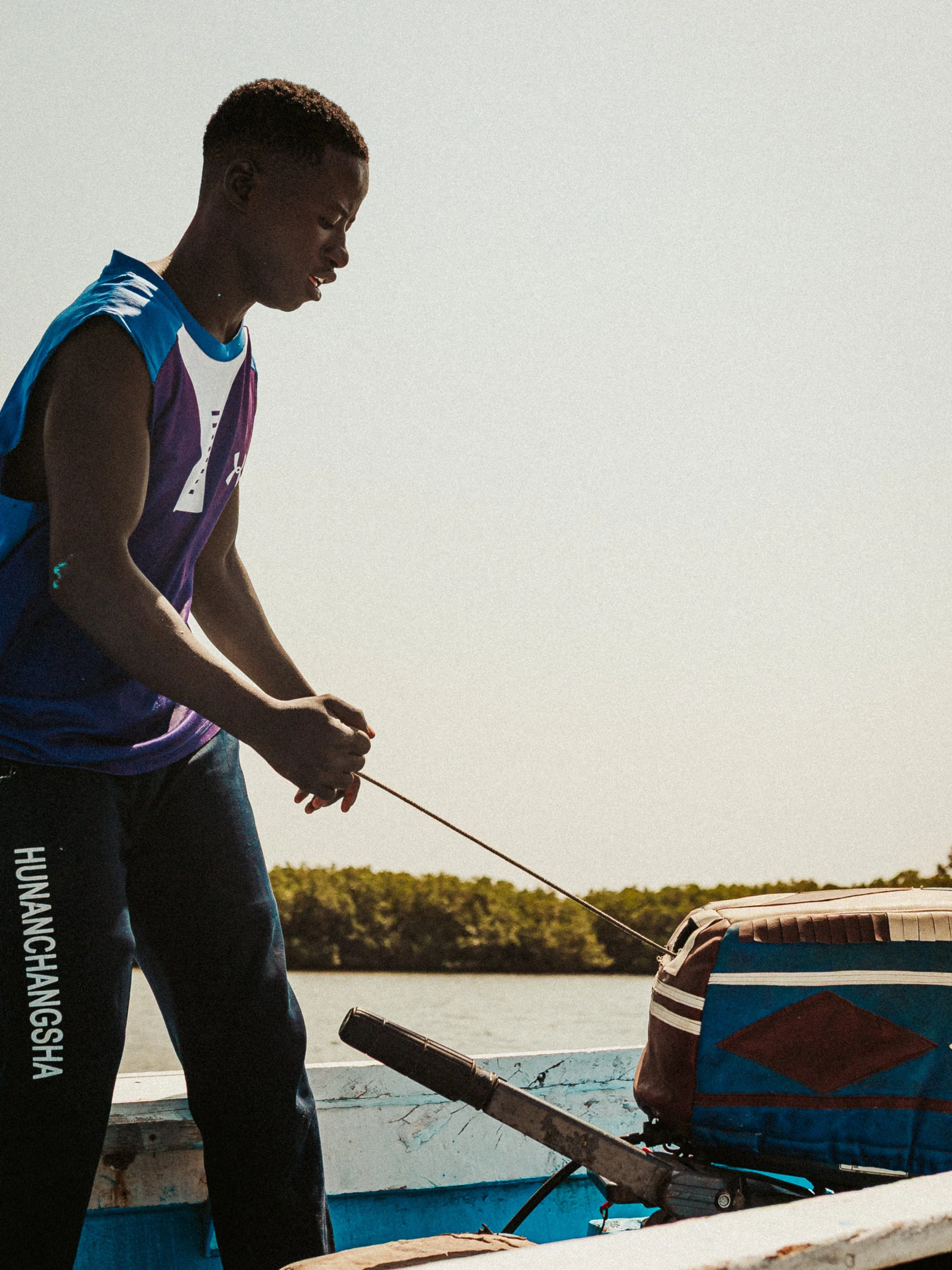 Jeune homme noir en T-shirt sans manches, tenant une corde ou câble, près d'une barque avec un sac de sport coloré, en plein air au bord d'une rivière ou lac, avec des arbres en arrière-plan, sous un ciel clair.