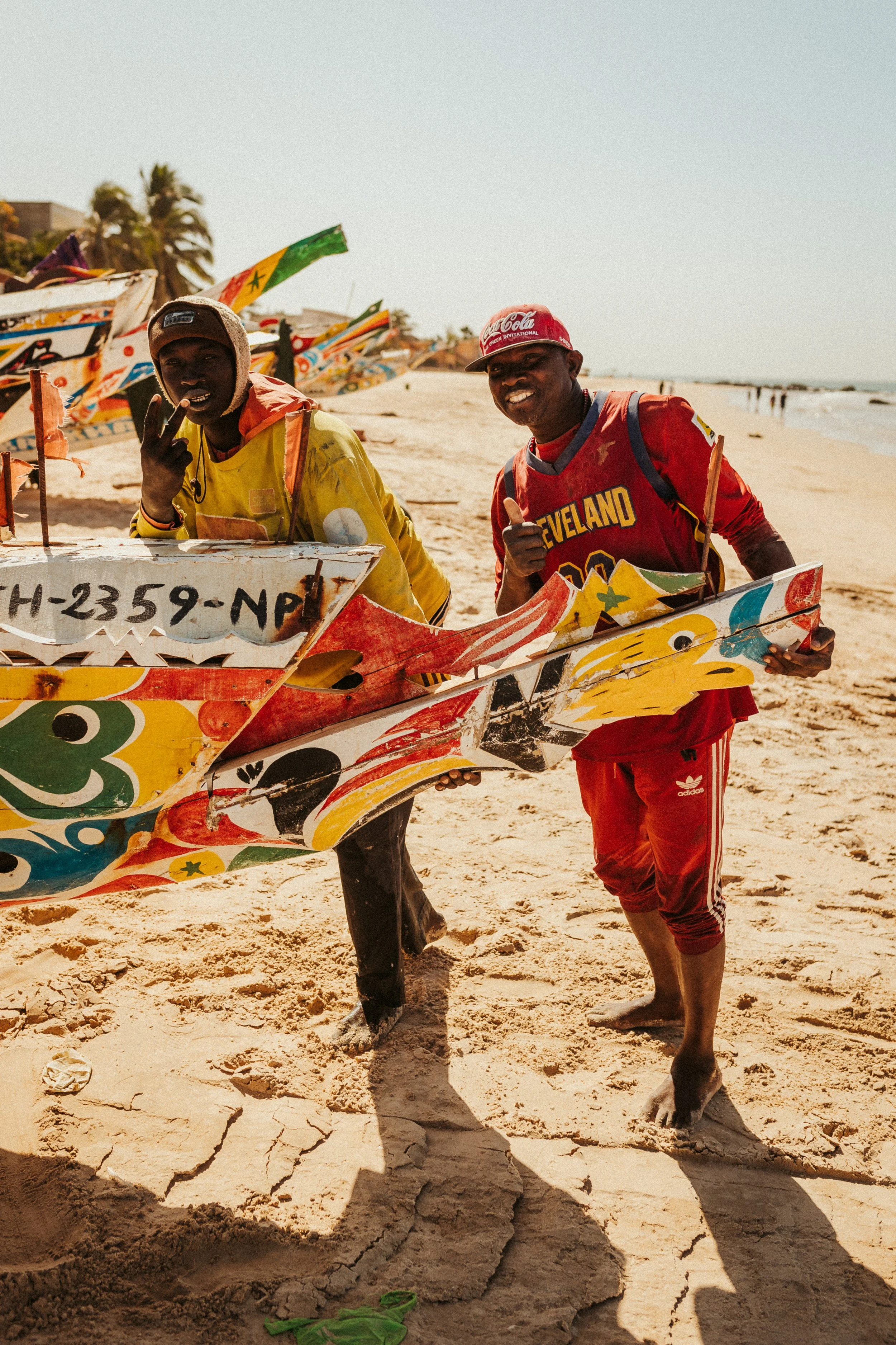 Deux hommes souriants posent avec une planche de surf colorée sur la plage ensoleillée, avec d'autres planches en arrière-plan.