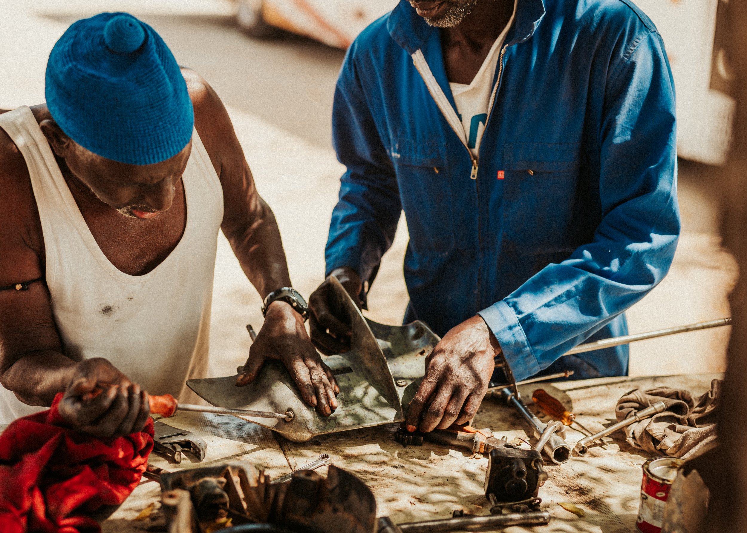 Deux hommes travaillent ensemble sur un moteur ou une pièce mécanique, avec divers outils et pièces autour d'eux, dans un environnement de bricolage ou d'atelier.