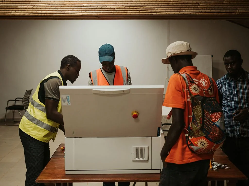 Quatre hommes regardent un appareil, probablement une machine à voter ou un équipement électoral, dans une salle intérieure, certains portant des gilets de sécurité ou des chapeaux, une activité semble en cours de vérification ou de démonstration.