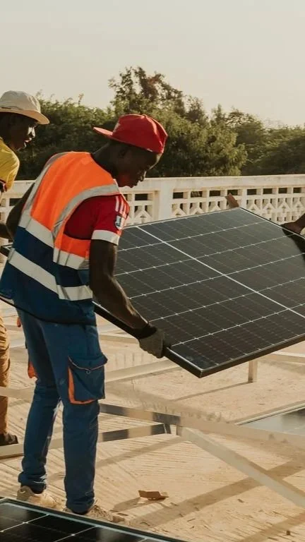 Deux personnes installant un panneau solaire sur un support en bois, avec un fond de végétation et de ciel clair.