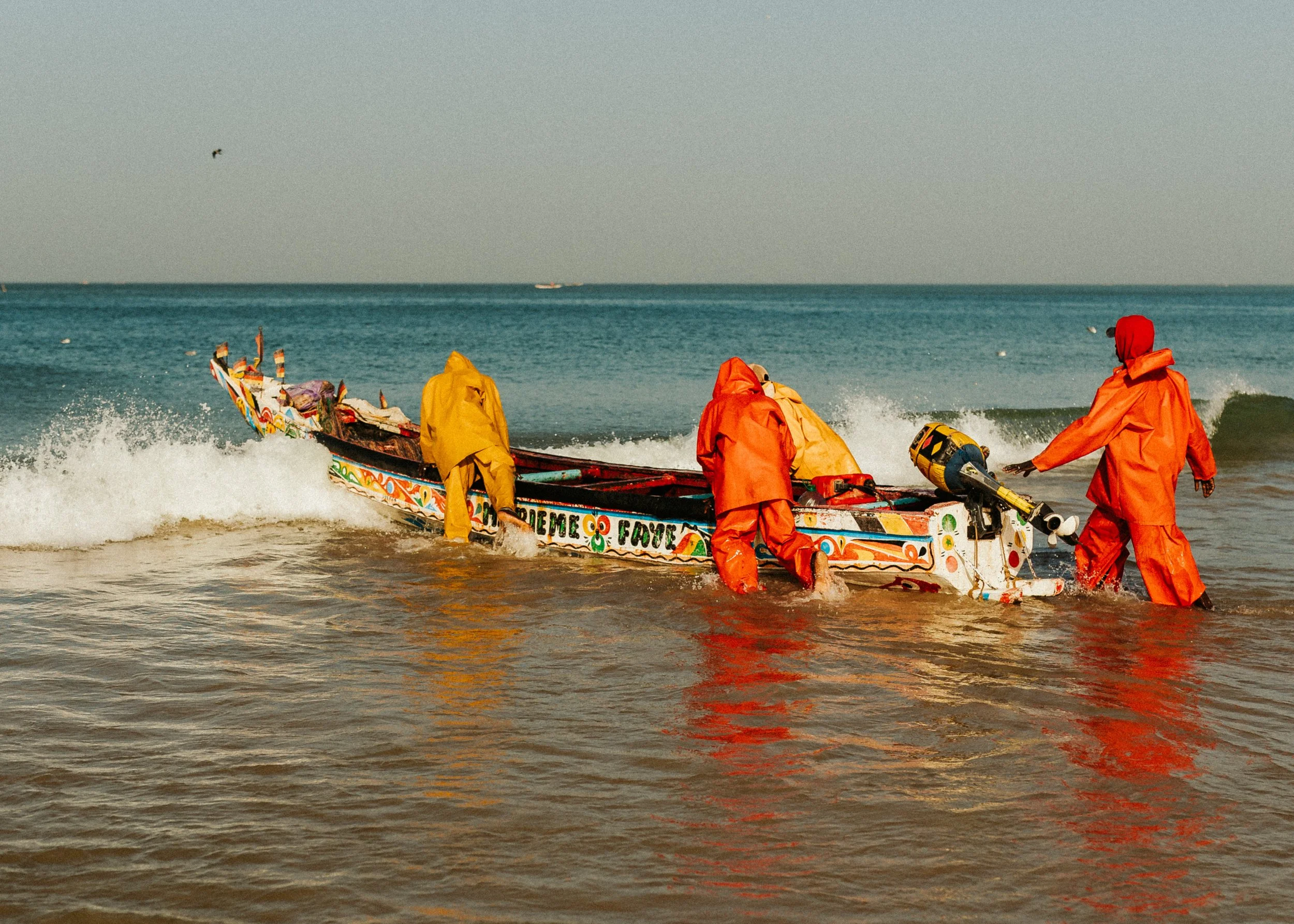 Personnes en tenue orange à la plage avec un bateau coloré au bord de l'eau.