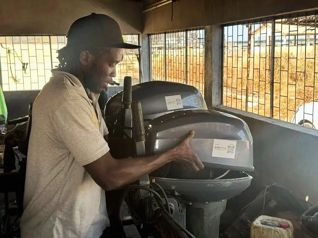 Un homme examine un moteur de bateau dans un atelier, avec des fenêtres en grille laissant entrer de la lumière naturelle.