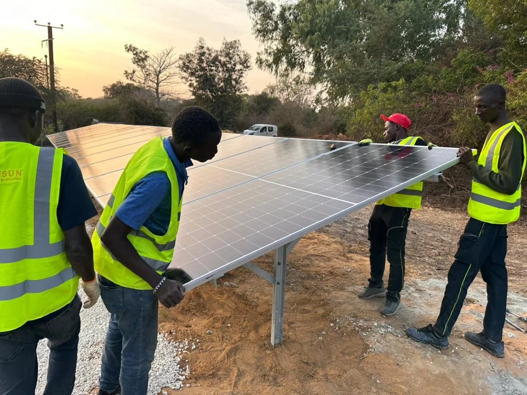 Cinq hommes en gilets de sécurité jaunes installant un panneau solaire photovoltaïque en extérieur, avec des arbres et un ciel au coucher du soleil en arrière-plan.