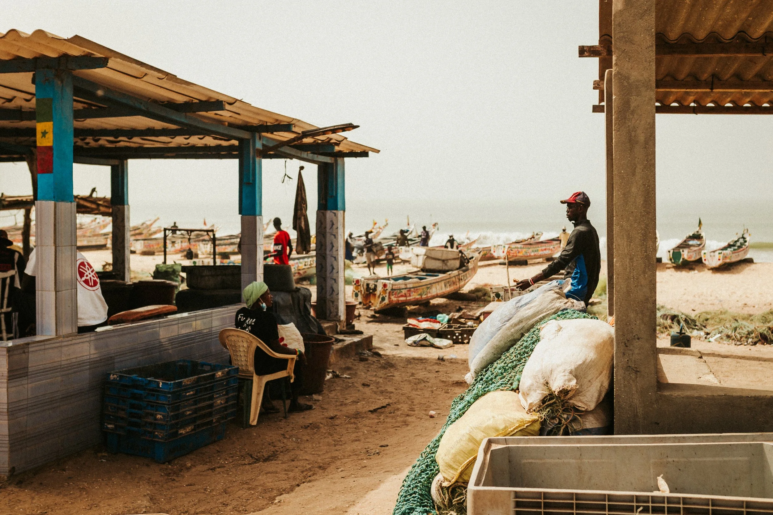 Marché de pêcheurs sur une plage avec des bateaux de pêche traditionnels en arrière-plan, plusieurs personnes assises ou discutant. Des sacs et des équipements de pêche sont visibles.