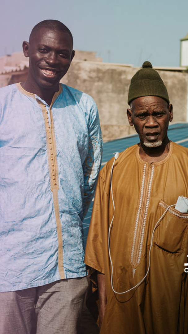 Deux hommes africains debout à l'extérieur, l'un souriant et l'autre avec une expression sérieuse, portant des vêtements traditionnels, avec un bâtiment en arrière-plan.