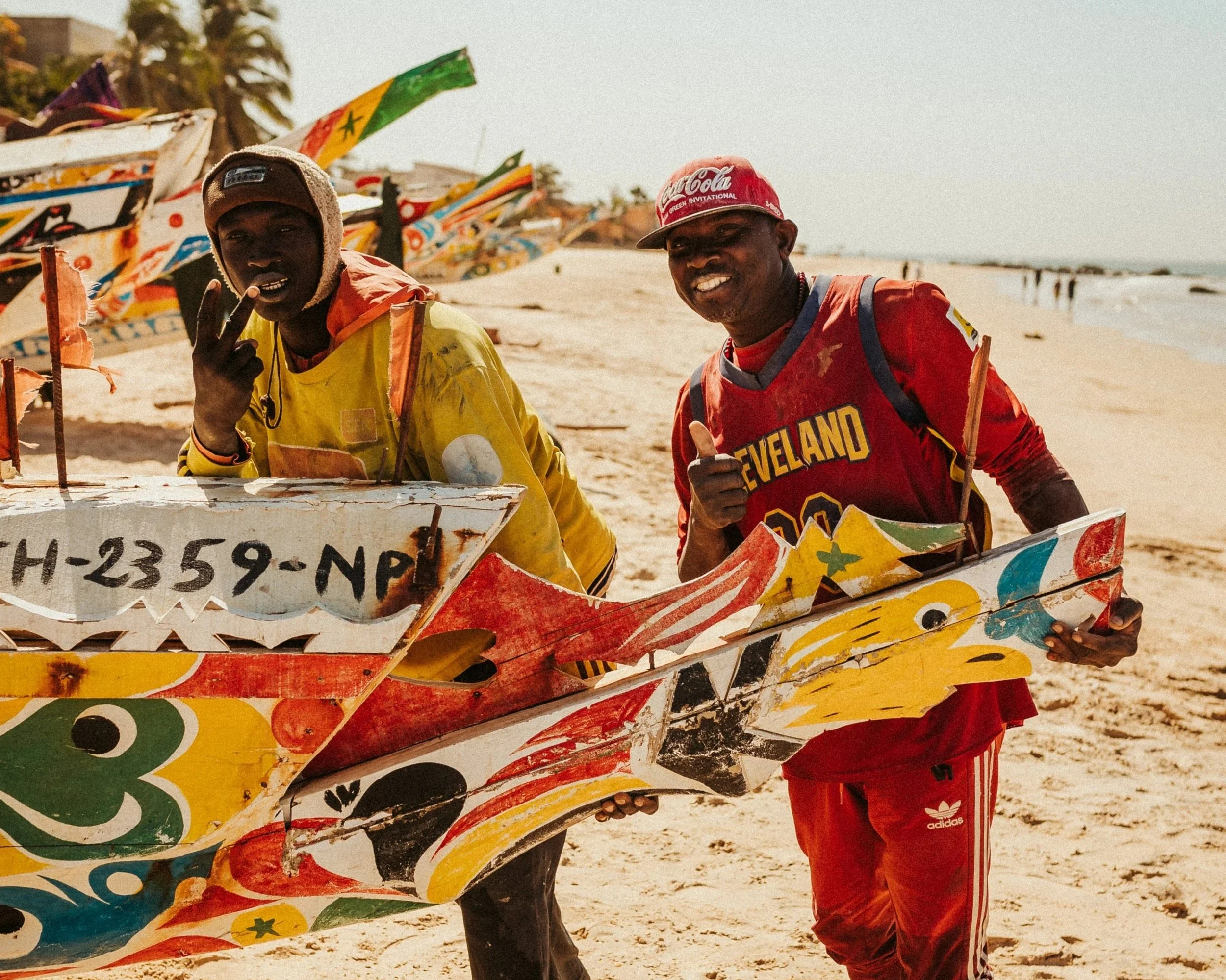 Deux hommes souriants avec des planches de surf peintes, en bord de plage. L'un porte un maillot de sport rouge avec 'Cleveland' et le numéro 00, l'autre une veste jaune, et ils font un signe de paix ou de victoire.