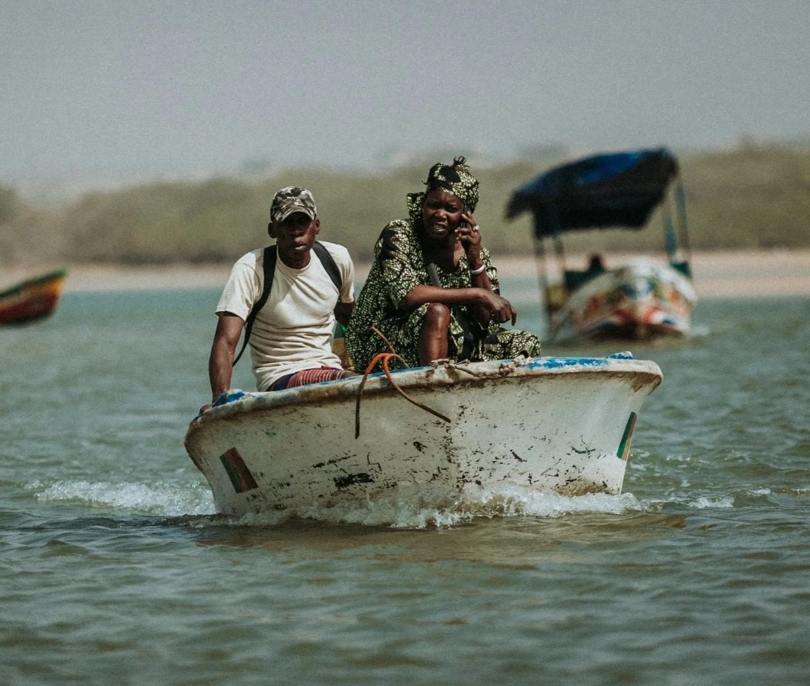 Une femme et un jeune homme sur un bateau au bord de l'eau, avec un bateau à moteur en arrière-plan, dans un paysage côtier.