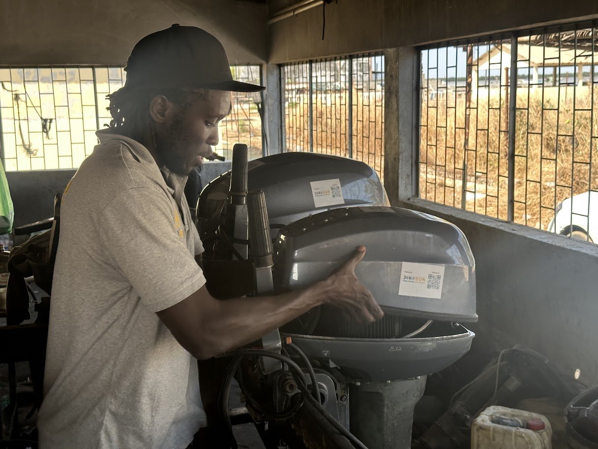 Un homme porte une casquette grise, travaille sur une machine à l'intérieur d'un bâtiment, avec des fenêtres en grillage décoratif et vue sur une zone extérieure ensoleillée.