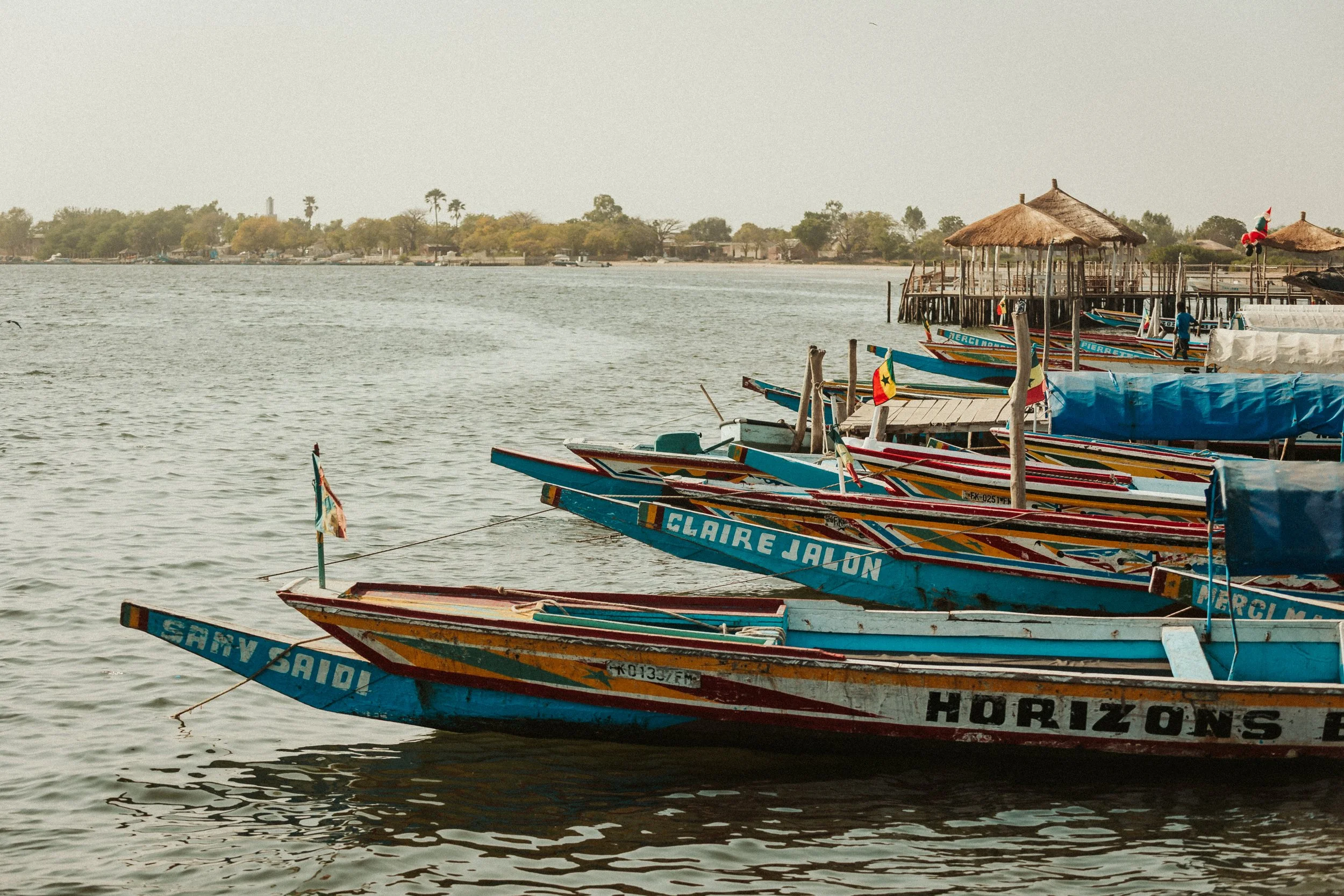 Bateaux de pêche colorés amarrés au quai et un village en arrière-plan sur la rive opposée, au bord d'une rivière ou d'un lac.