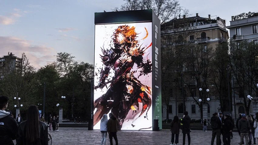 Un grande schermo digitale che mostra un'illustrazione astratta di un personaggio fantasy con armatura e arma, in un parco urbano nel centro di Milano, durante la sera. L'opera digitale si chiama Vortex di Marcello Baldari.